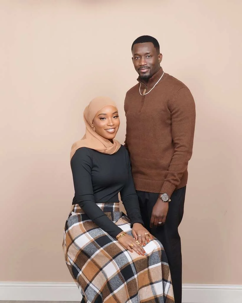 A woman wearing a beige hijab and dark top, sitting, and a man in a brown sweater and black pants standing beside her against a plain beige background.