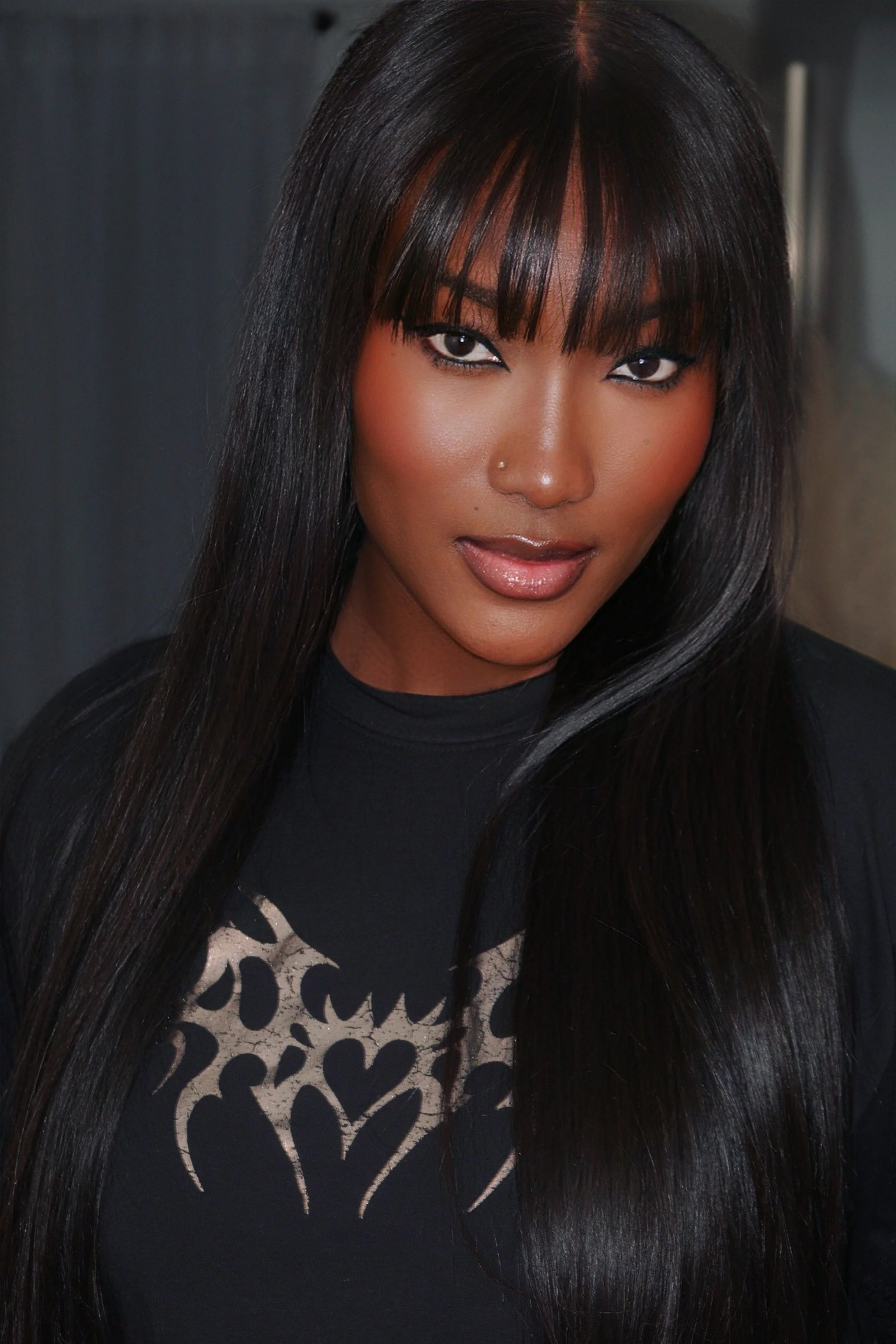 A woman with long, straight black hair and bangs, wearing makeup and a nose piercing, posing indoors in a black top with a paisley design.