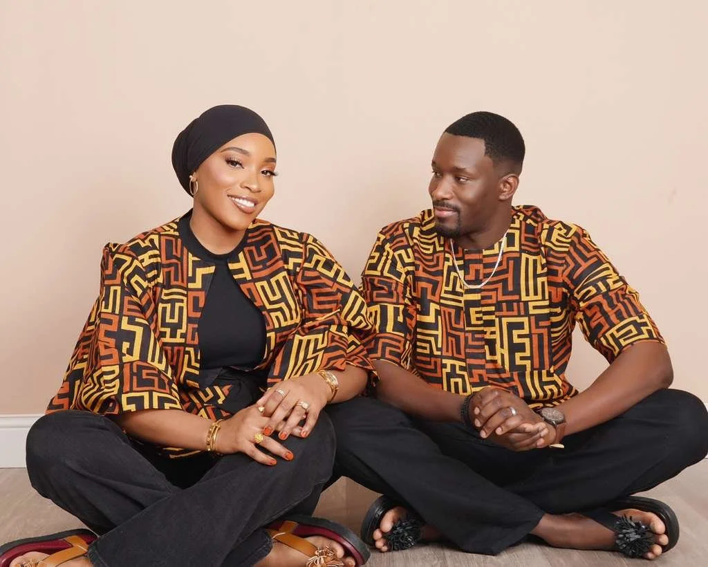 A smiling woman wearing a black headscarf, black top, and patterned jacket, sitting cross-legged on the floor next to a man with short hair, wearing a matching patterned shirt, black pants, and sandals, both sitting against a plain beige wall.