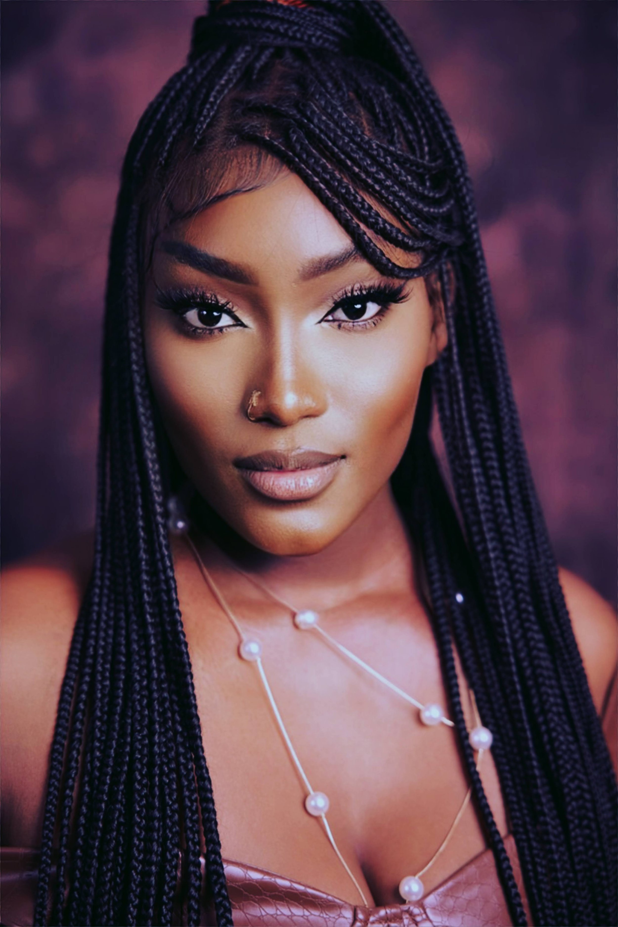 A woman with dark braided hair and gold nose ring looking directly at the camera, wearing pearl jewelry, with a blurred purple background.