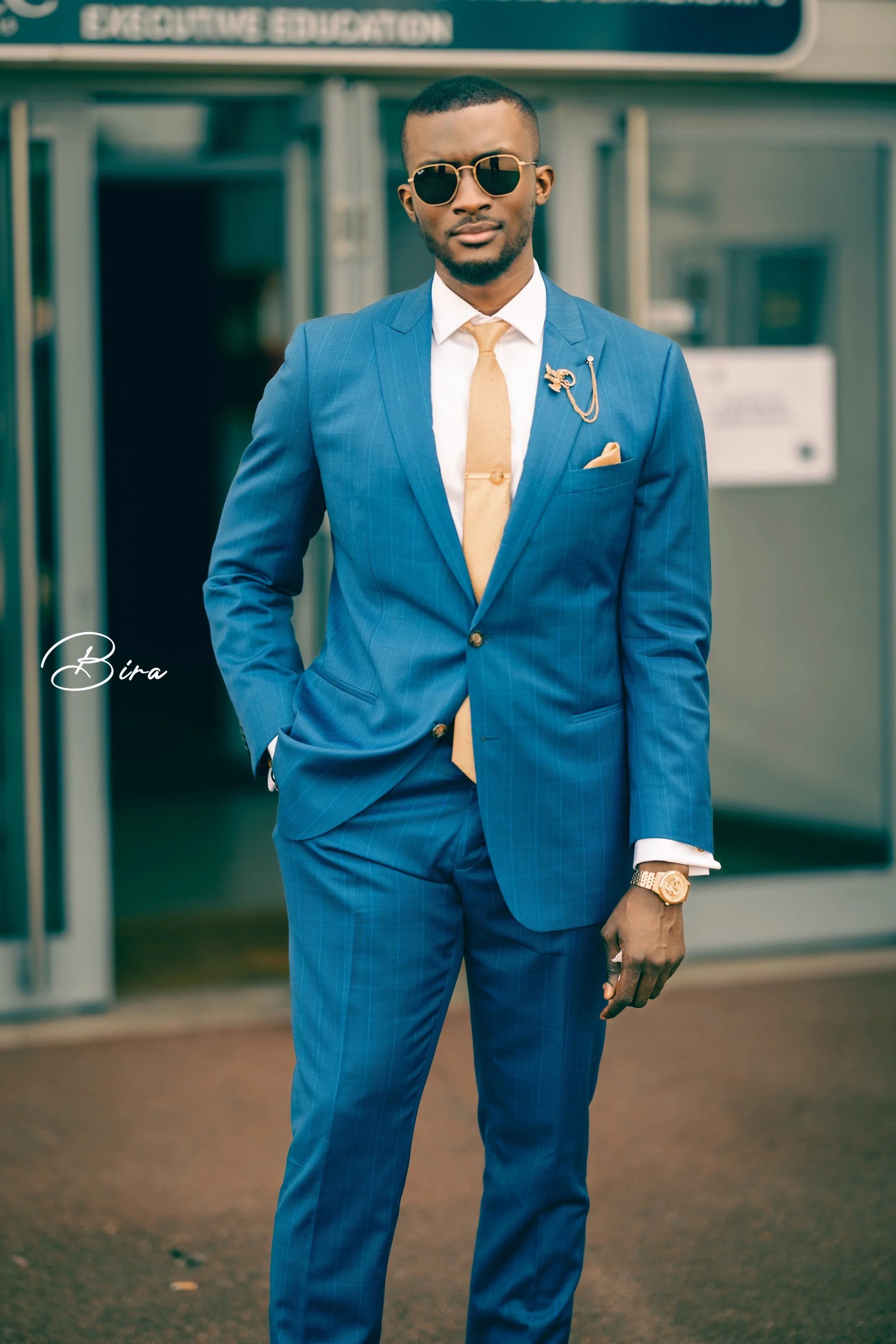 A man in a blue suit with a beige tie, white shirt, sunglasses, and a gold watch, standing outside a building with glass doors, with his right hand in his pocket and left hand resting by his side.