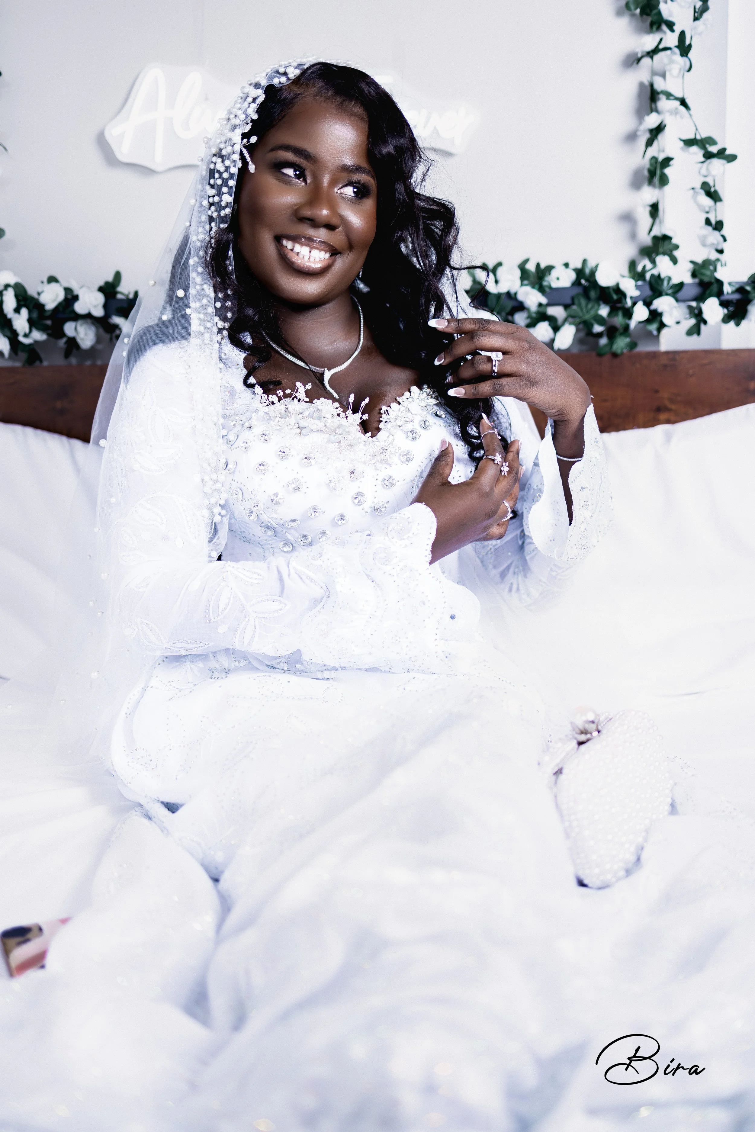 A woman in a white wedding dress is sitting on a bed, smiling and touching her chest. She wears a veil, necklace, and rings, with a floral headpiece. Decor and a sign are visible in the background.