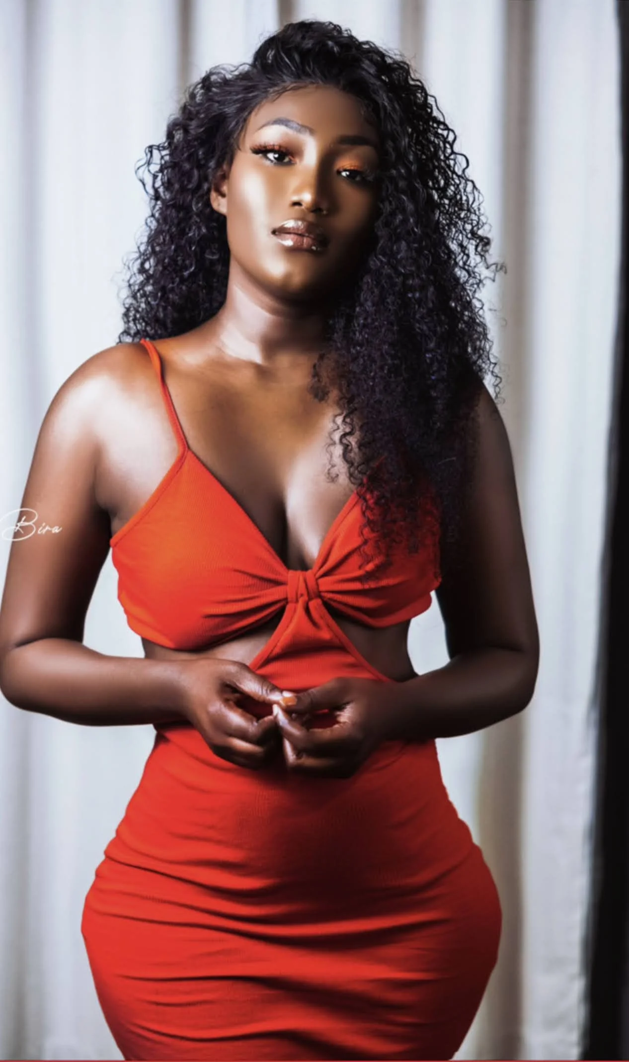 A woman with dark, curly hair wearing a red dress with a knotted front, standing indoors in front of a light-colored curtain.