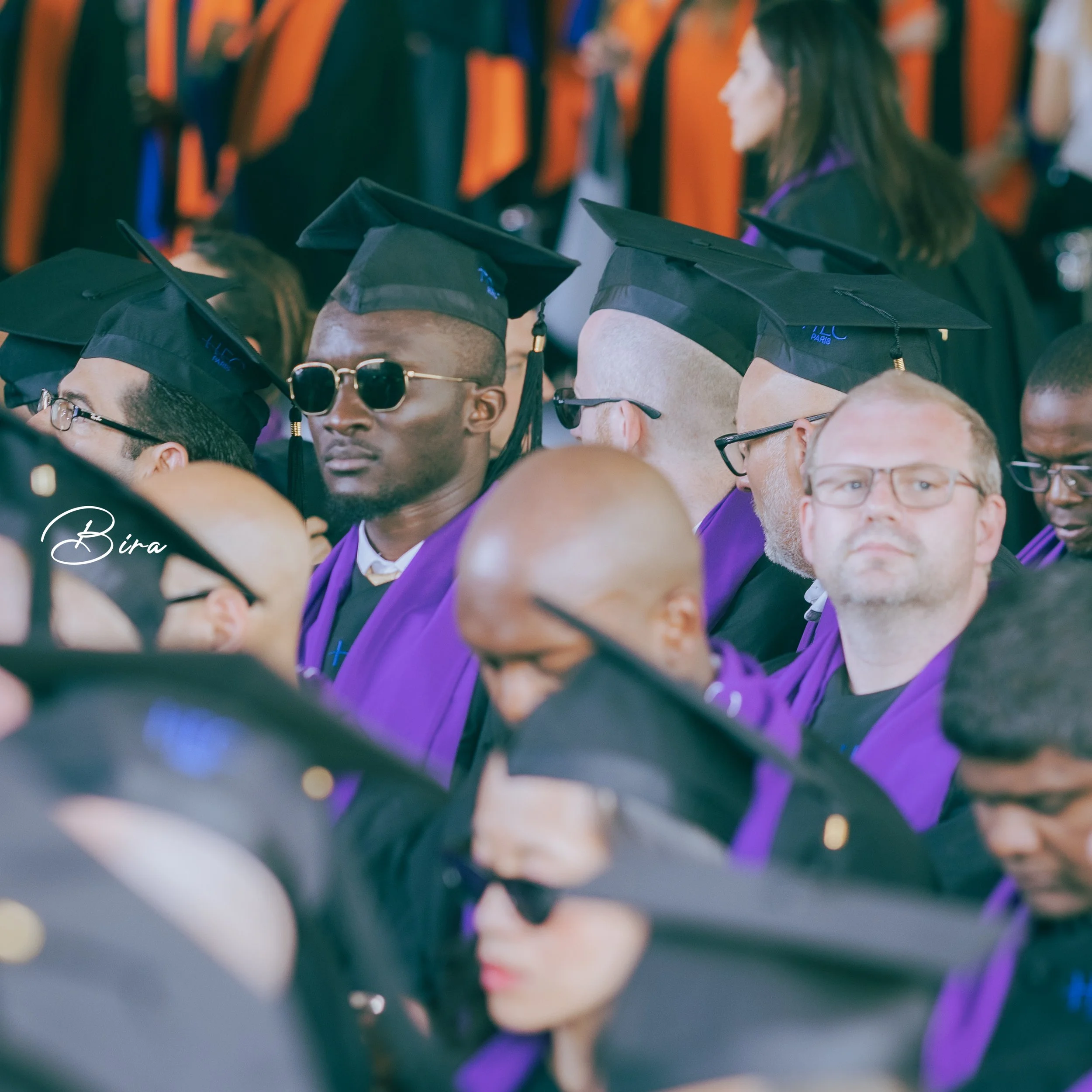 Graduates in caps and gowns seated during a graduation ceremony, some wearing sunglasses, with a mostly focus on a man with dark glasses in the center.