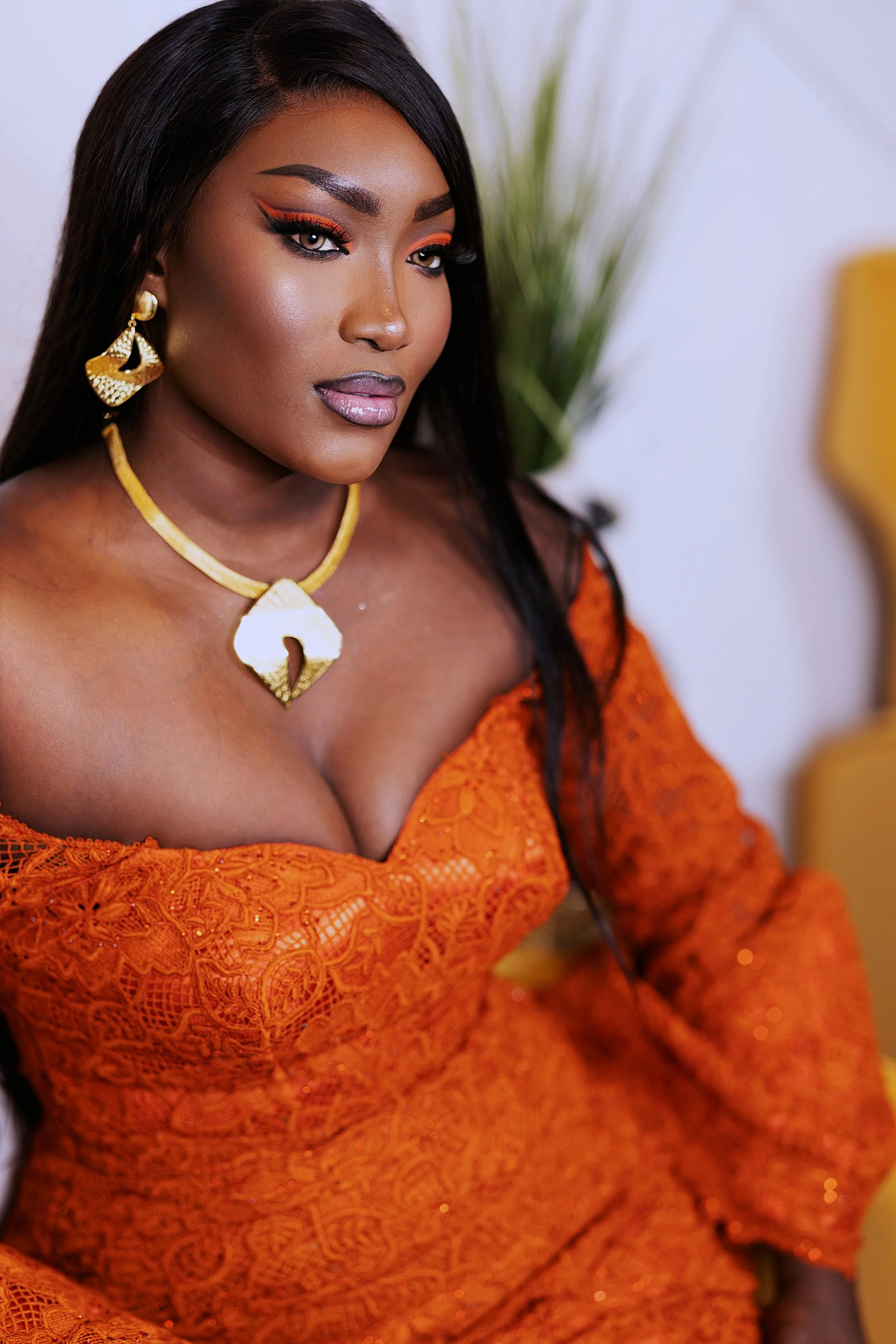 A woman with long dark hair and makeup, wearing orange lace clothing and gold jewelry, sitting indoors with a plant and a yellow chair in the background.