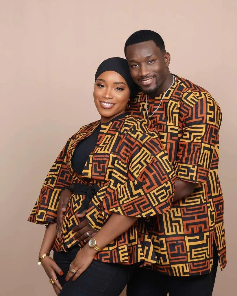 A smiling couple, a woman wearing a black headscarf and patterned top, and a man in matching patterned attire, standing close together against a plain beige background.