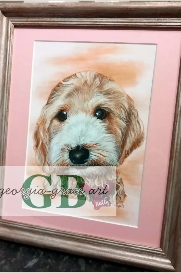 A framed watercolor painting of a puppy with a pink mat background, the puppy has light brown, white, and tan fur, with dark eyes and nose.