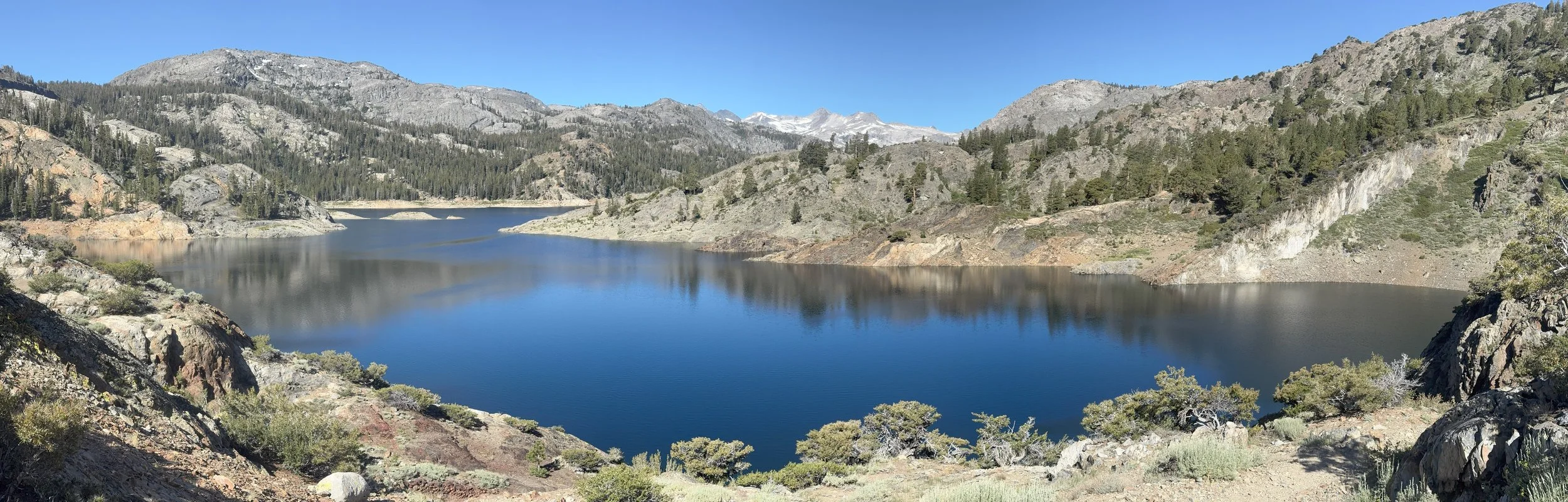 Scenic view of a calm mountain lake surrounded by rocky and forested hills under a clear blue sky.