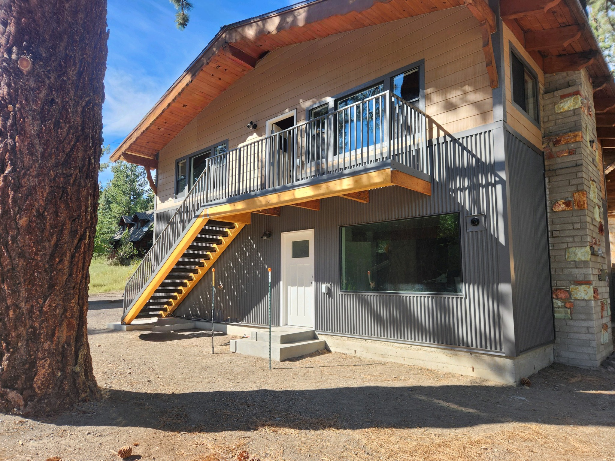 A modern two-story house with an exterior staircase, metal railing, and a balcony, surrounded by trees on a sunny day.