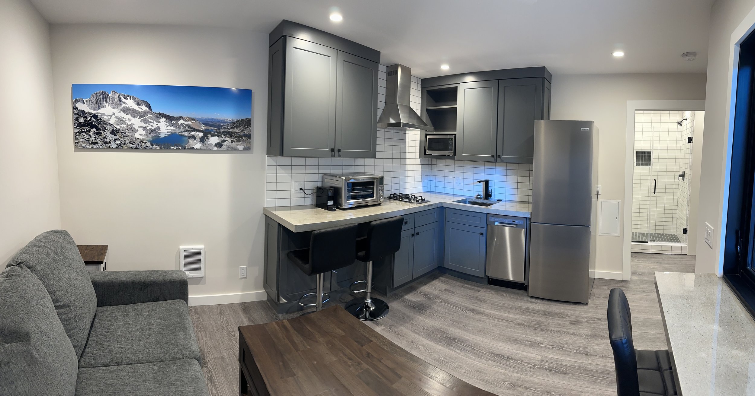 Open-concept living area with kitchen, gray cabinets, white tile backsplash, stainless steel appliances, a gray couch, a wooden coffee table, and a panoramic mountain landscape photograph on the wall.