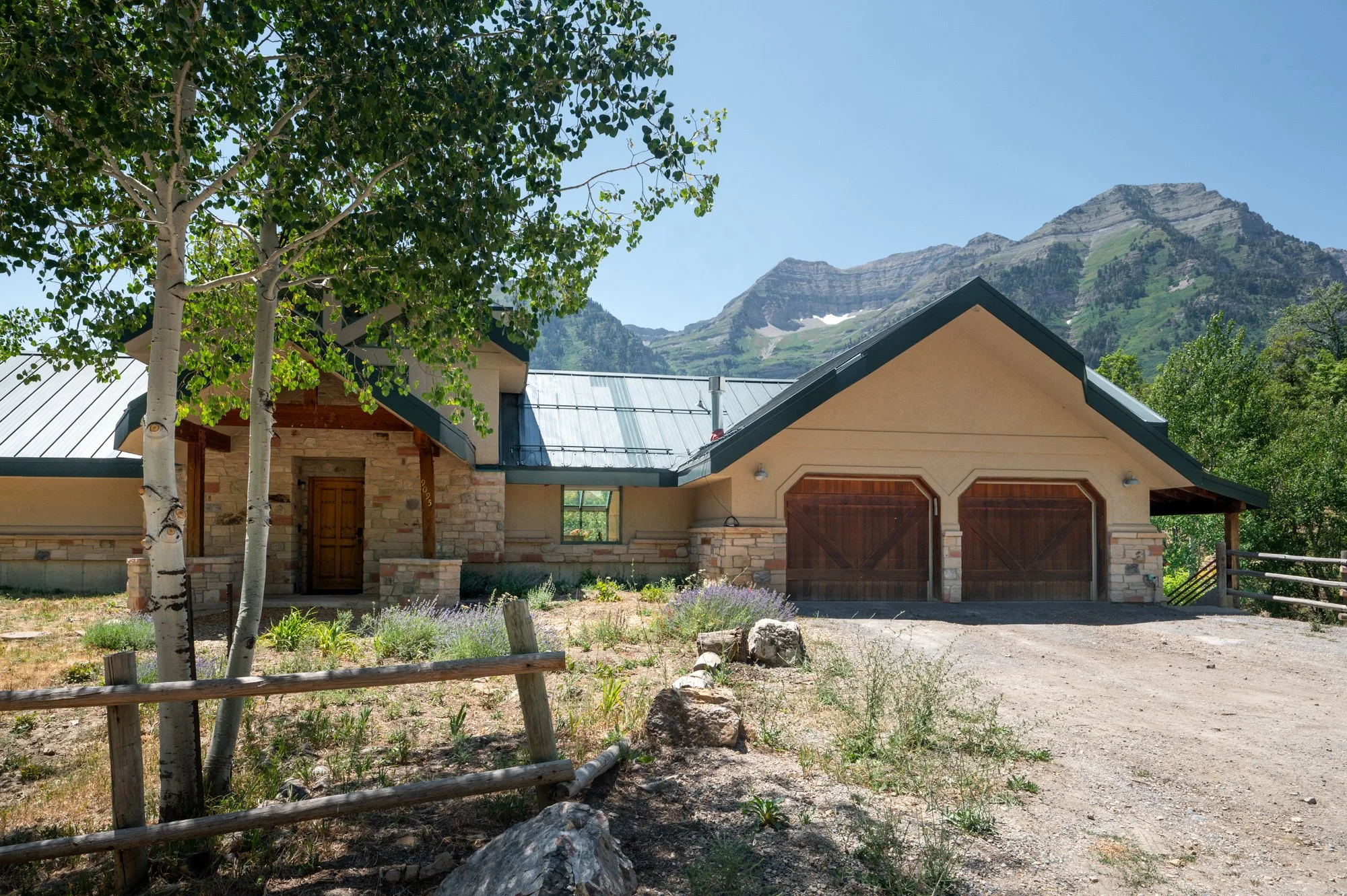 A house with stone and beige siding, wooden garage doors, and a metal roof, surrounded by trees, rocks, and mountainous terrain in the background.