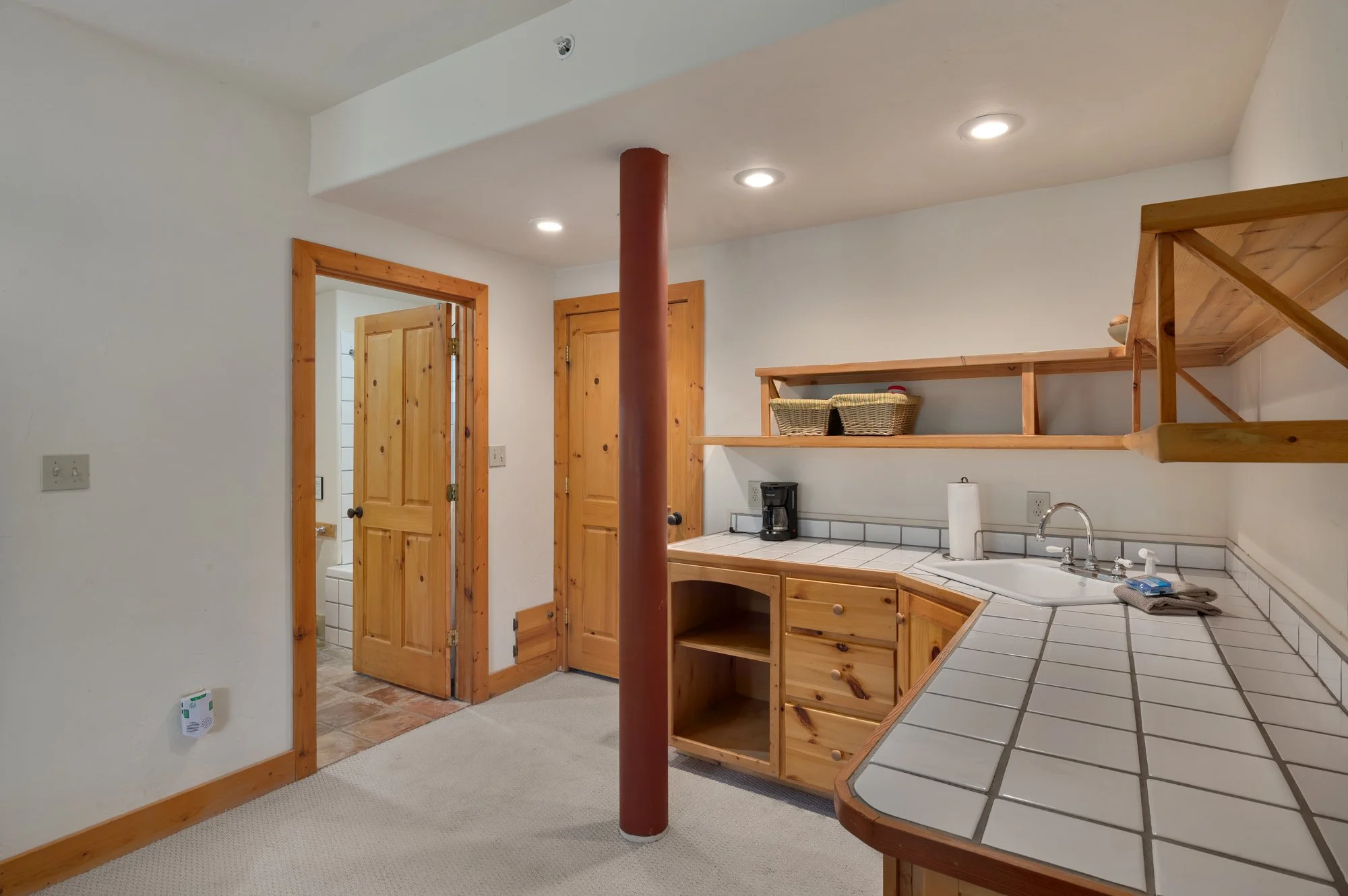 Interior view of a rustic kitchen with wooden cabinets, open shelves, and a tiled countertop. There is a coffee maker, paper towel, and a small sink. A red support pole stands in the middle, and a doorway leads to a bathroom with a toilet.