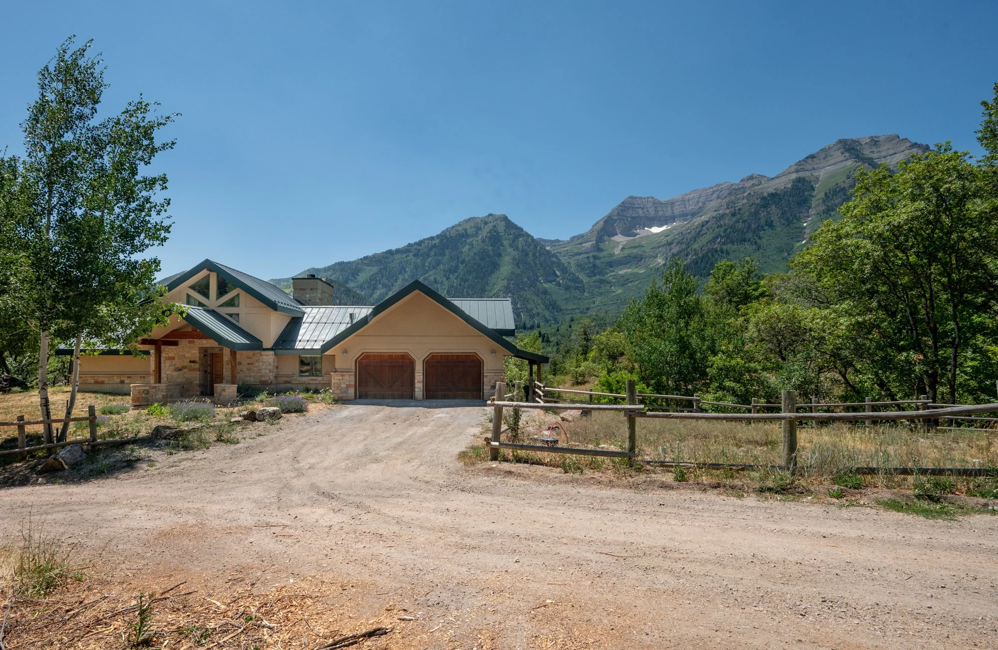 A modern house with a combination of stone and wood exterior, metal roof, facing a dirt driveway, surrounded by green trees, with mountains in the background and a clear blue sky overhead.