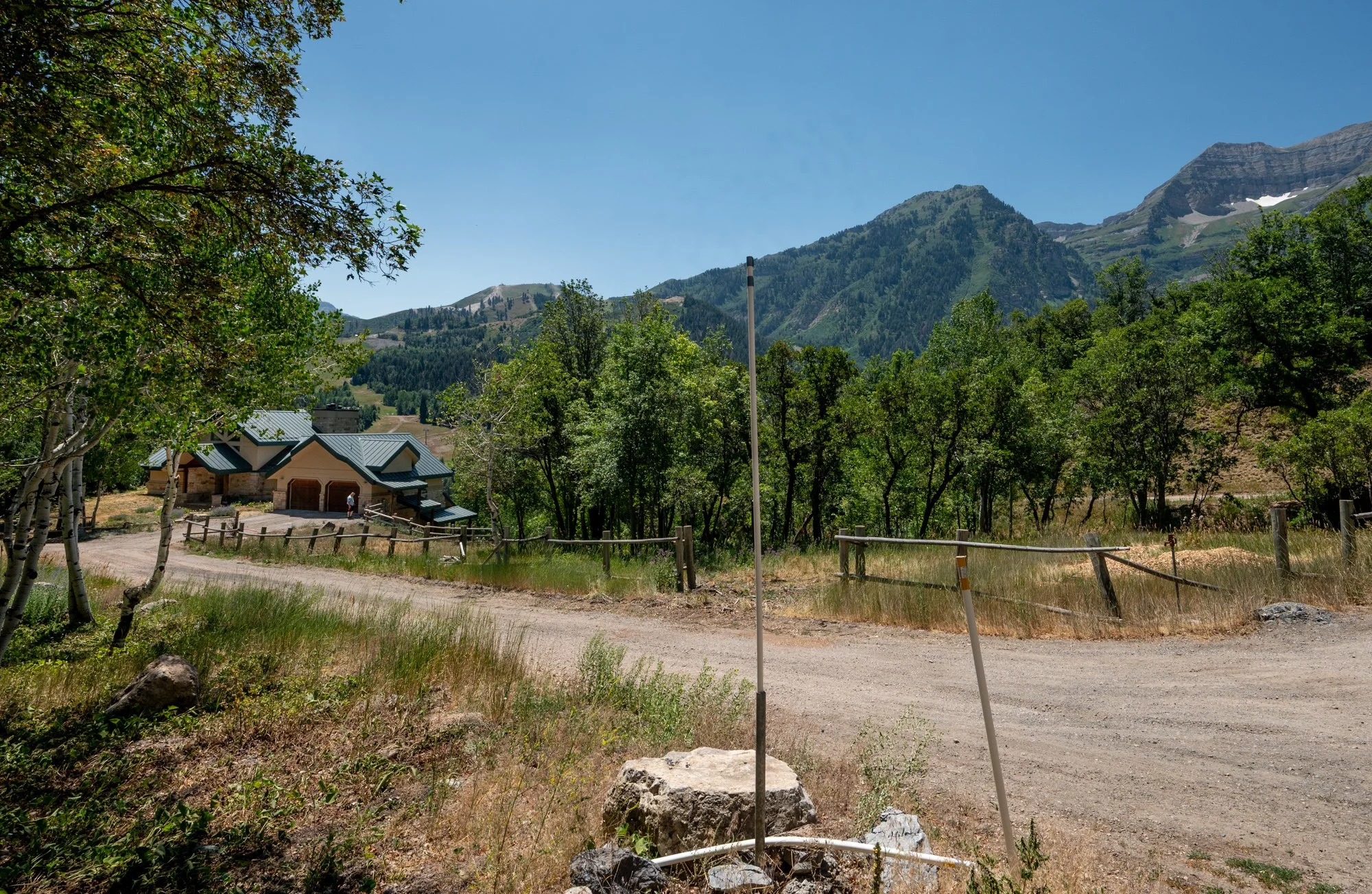 A dirt road leading to a house with green rooftops in a mountainous area, surrounded by trees and forested hills under a clear blue sky.
