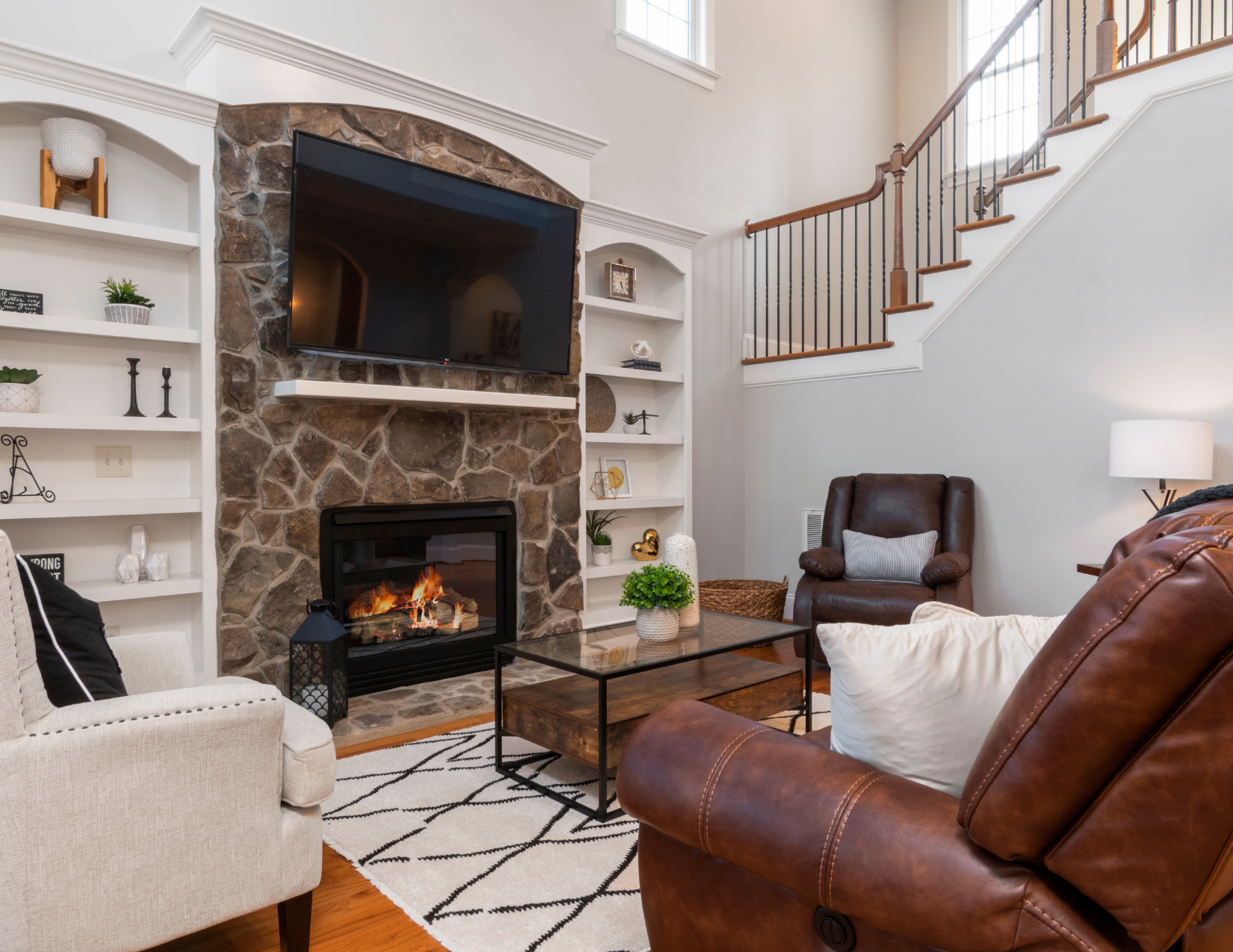 Living room with a stone fireplace, wall-mounted TV, white built-in shelves, and brown leather and beige upholstered armchairs, coffee table, and decorative lamps.