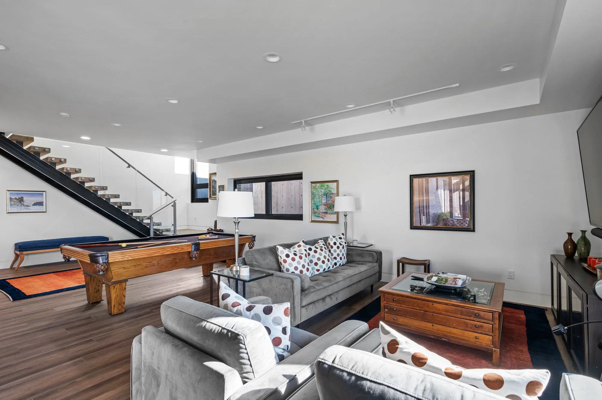 Living room with gray sofa, armchair, wooden coffee table, and a pool table. Walls decorated with framed artwork, windows letting in natural light, and a staircase in the background.