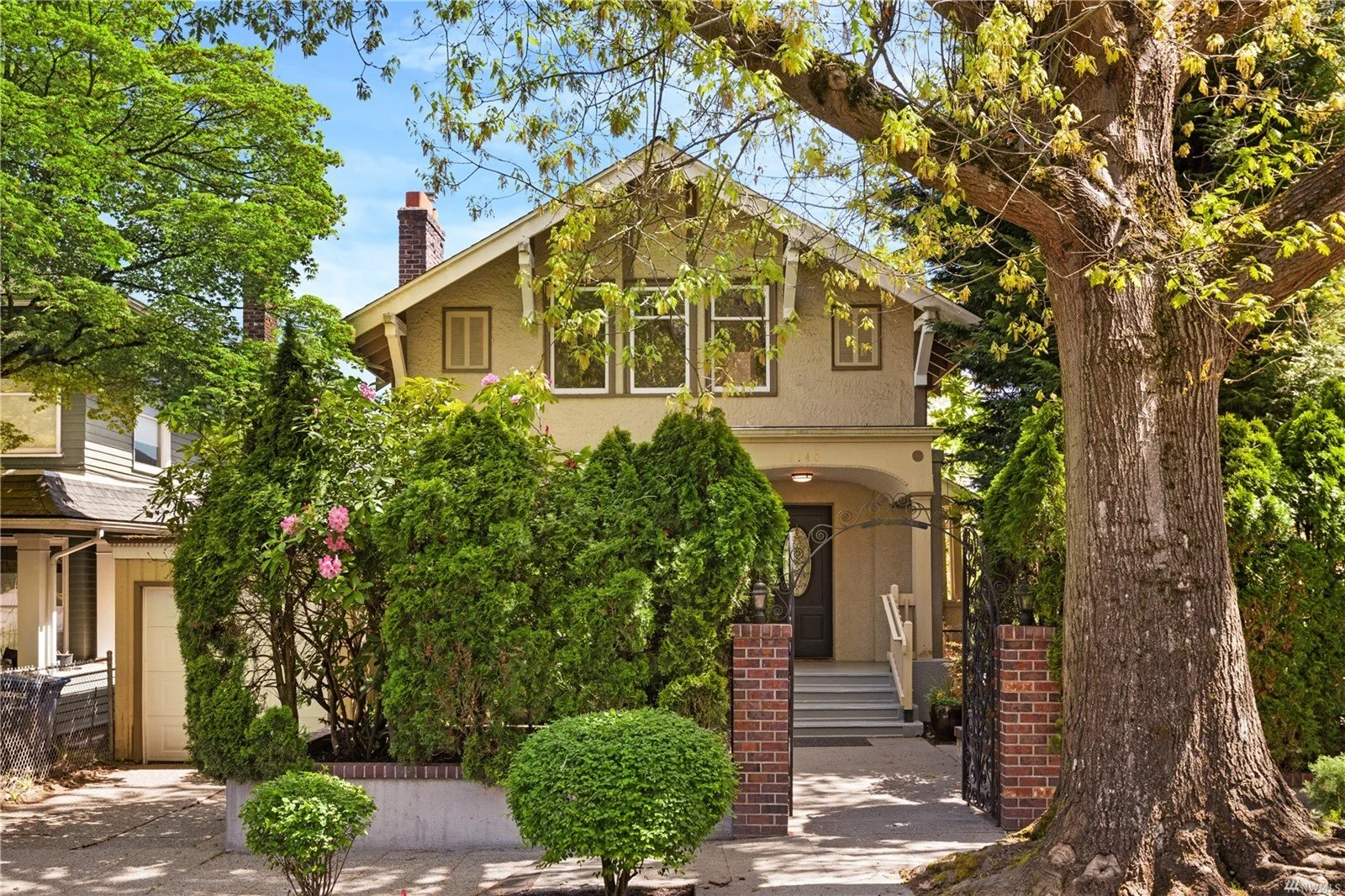 Front view of a two-story house with beige stucco exterior, surrounded by trees and greenery, with a brick post gate, and a large tree on the right.