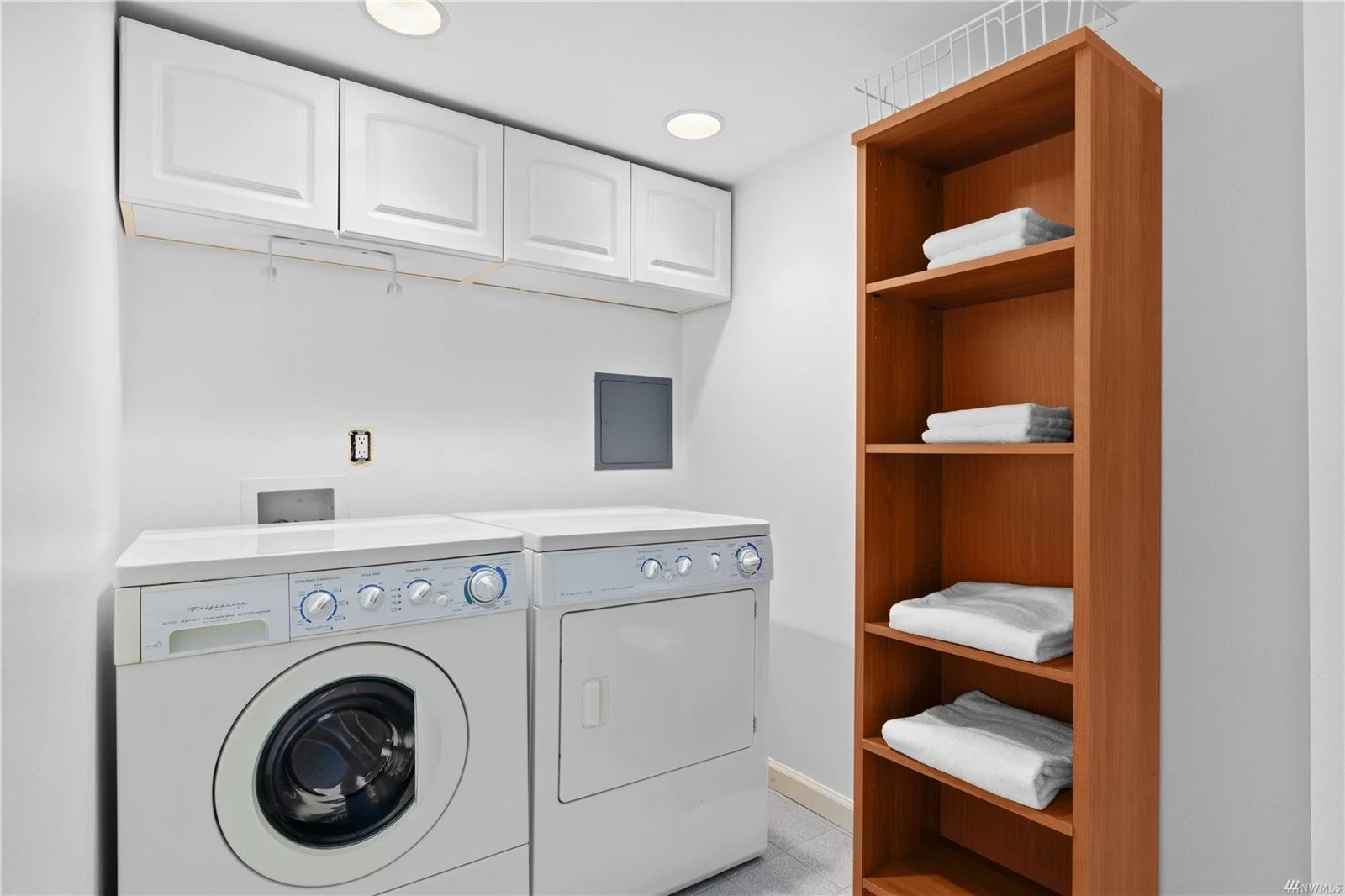 Laundry room with washing machine and dryer, white cabinets above, wooden shelving with folded towels, and white walls.