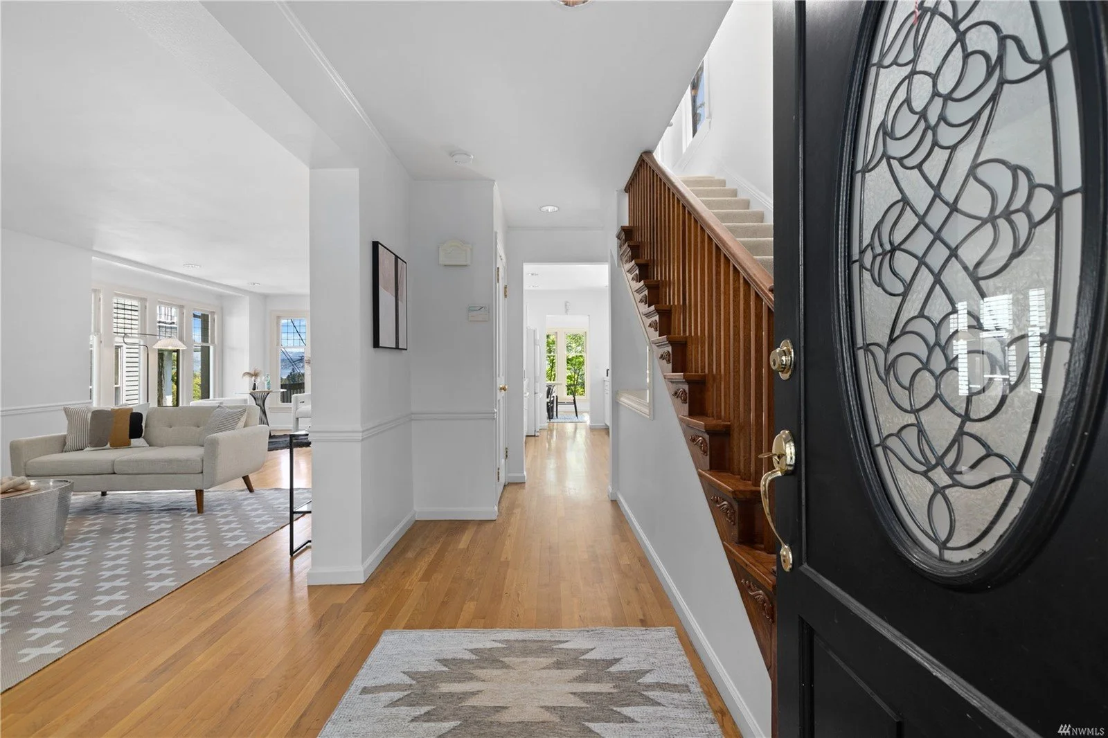 Inside the house showing a hallway with hardwood flooring, a wooden staircase, and a front door with decorative glass. The hallway leads to a bright living room with large windows and white walls.