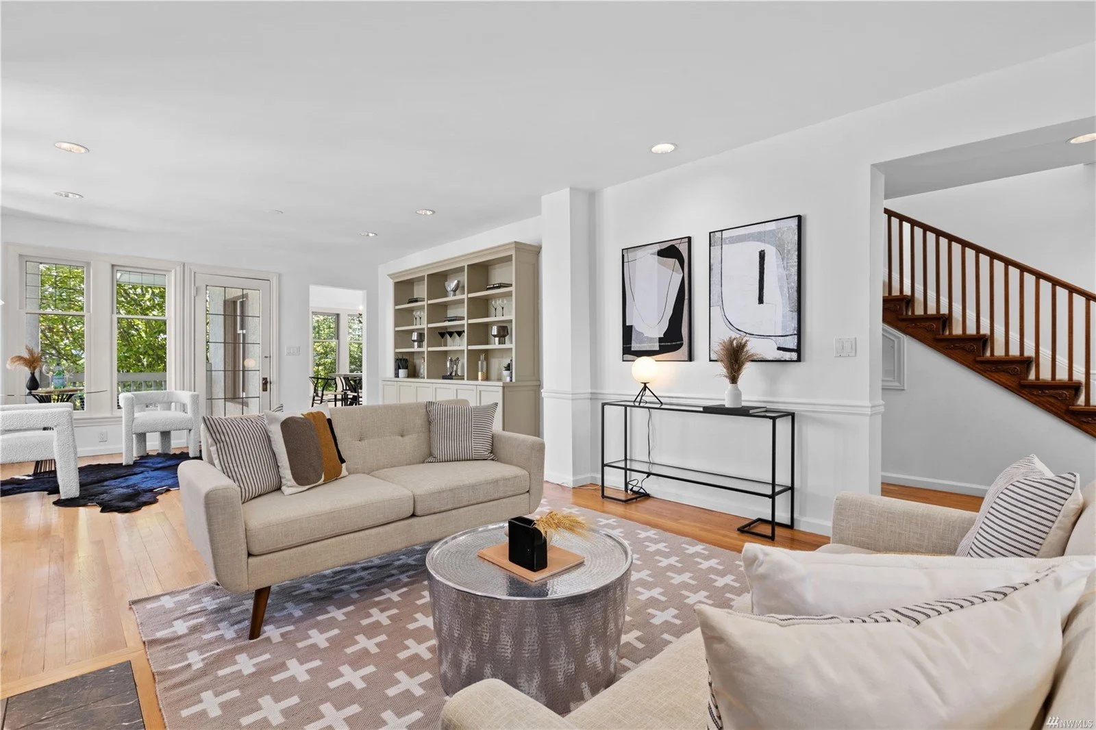 Bright living room with beige couches, patterned pillows, a round coffee table, abstract wall art, and a staircase with wooden railing.