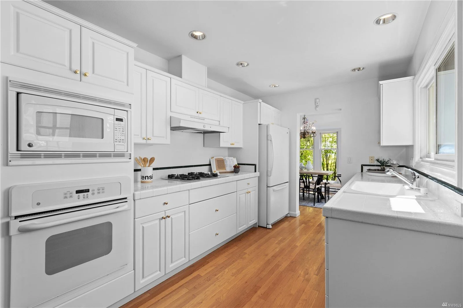 Clean white kitchen with wooden flooring, featuring white cabinets, a microwave and oven stacked together, a refrigerator, a gas stove, and a double-basin sink under a large window.