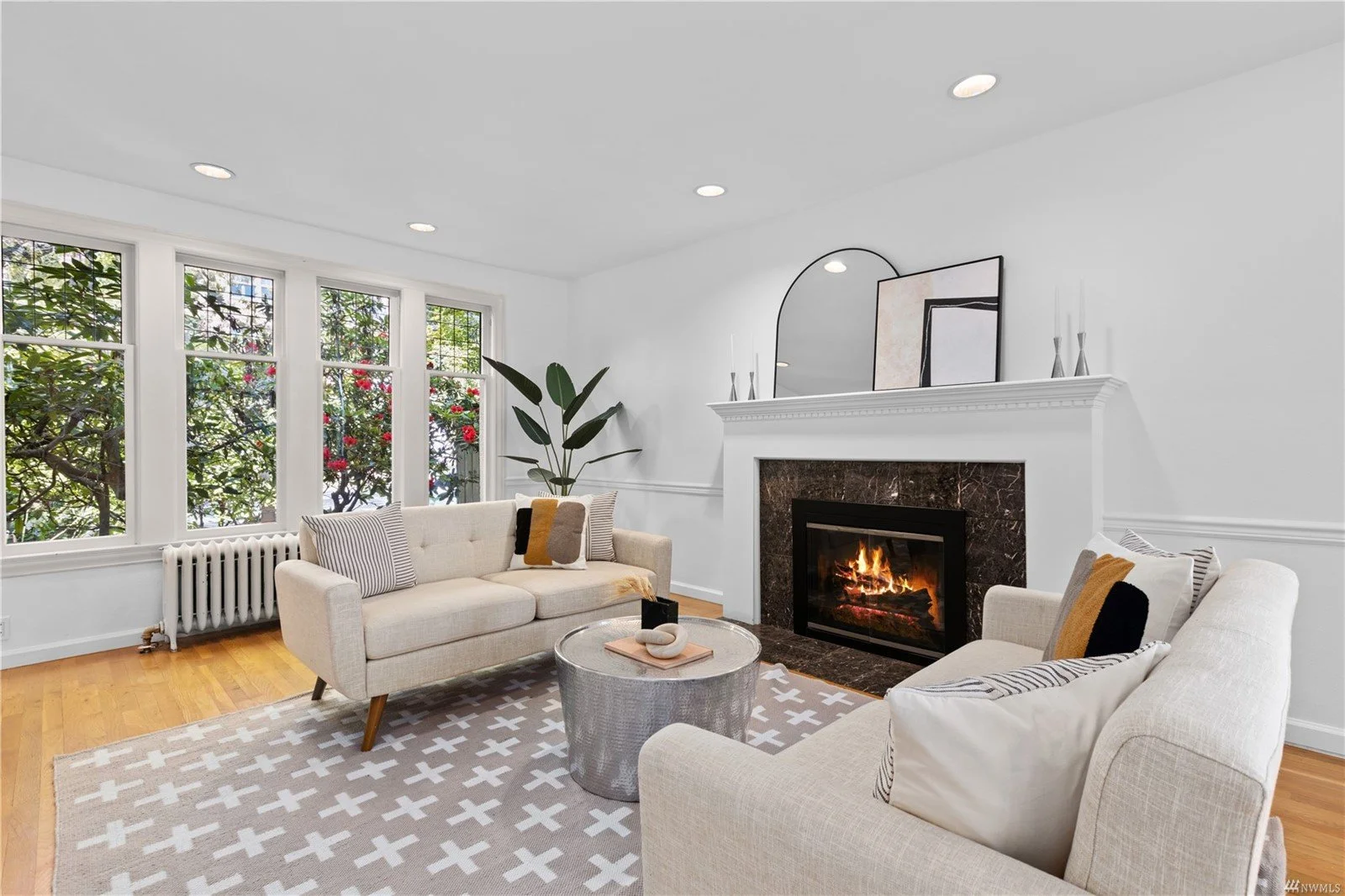 Living room with white walls, large framed windows with a view of greenery, a white sofa and a matching armchair with striped and solid colored pillows, a round coffee table, modern fireplace, decorative artwork, mirror, and a large plant in the corn