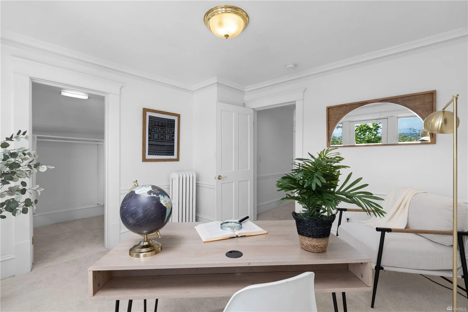 A bright and airy room with white walls and a ceiling light fixture. Features include an open closet, a wooden desk with a globe, an open book, and a magnifying glass, a potted green plant, a white sofa with a blanket, a gold floor lamp, and a decora