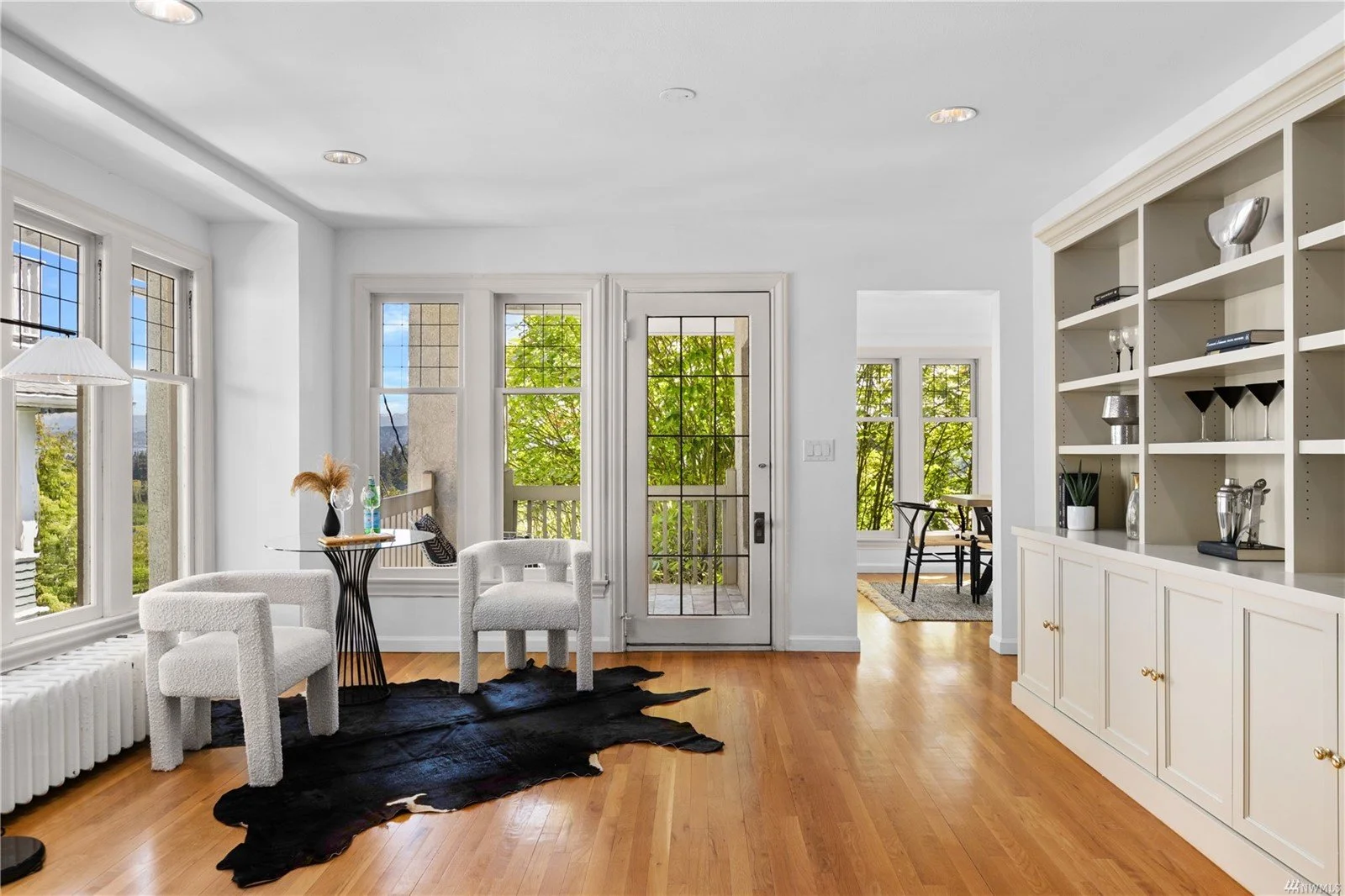 Bright living room with large windows, white walls, wooden flooring, two white upholstered chairs, a black animal hide rug, a small glass-top table with a decorative vase and bottle, a set of glassware and decor on a white built-in shelf, and a glass