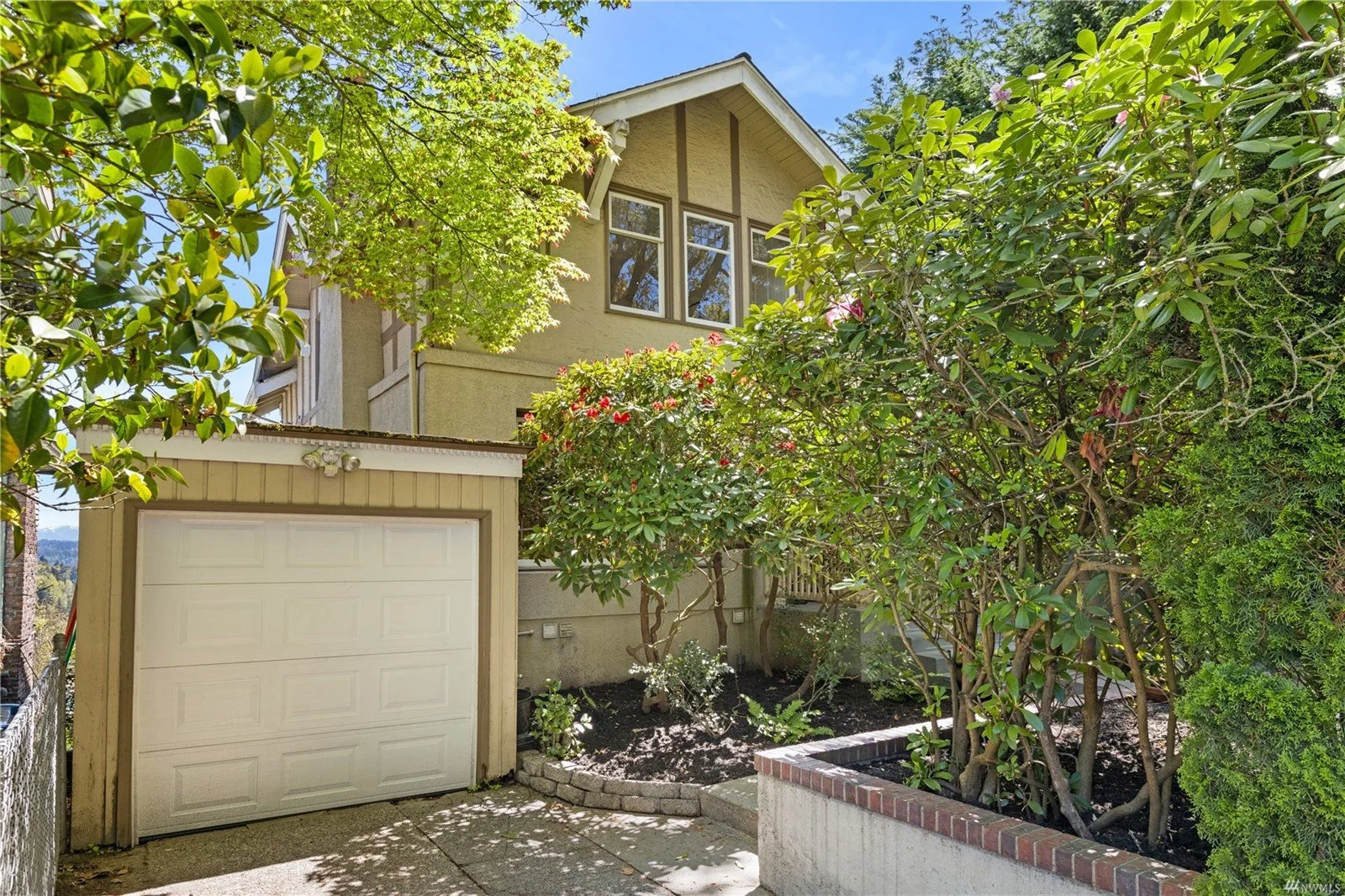Residential house with beige exterior, attached garage, surrounded by green trees and shrubs, with a small front yard and a brick planter wall.