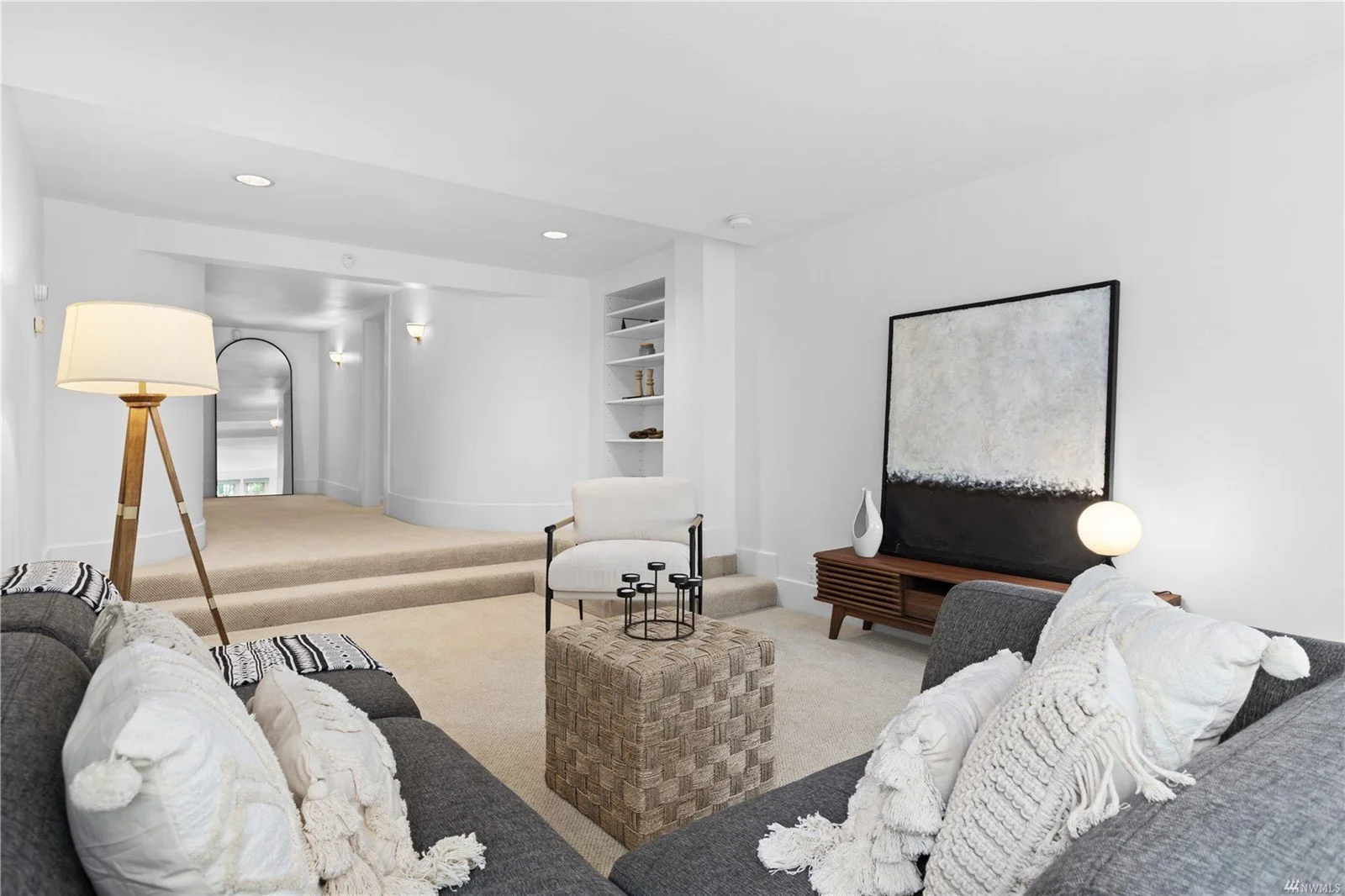 Living room with gray sofas, white pillows, a woven ottoman, a tall wooden floor lamp, a white armchair, and a wooden media console with a large abstract painting on the wall behind it.