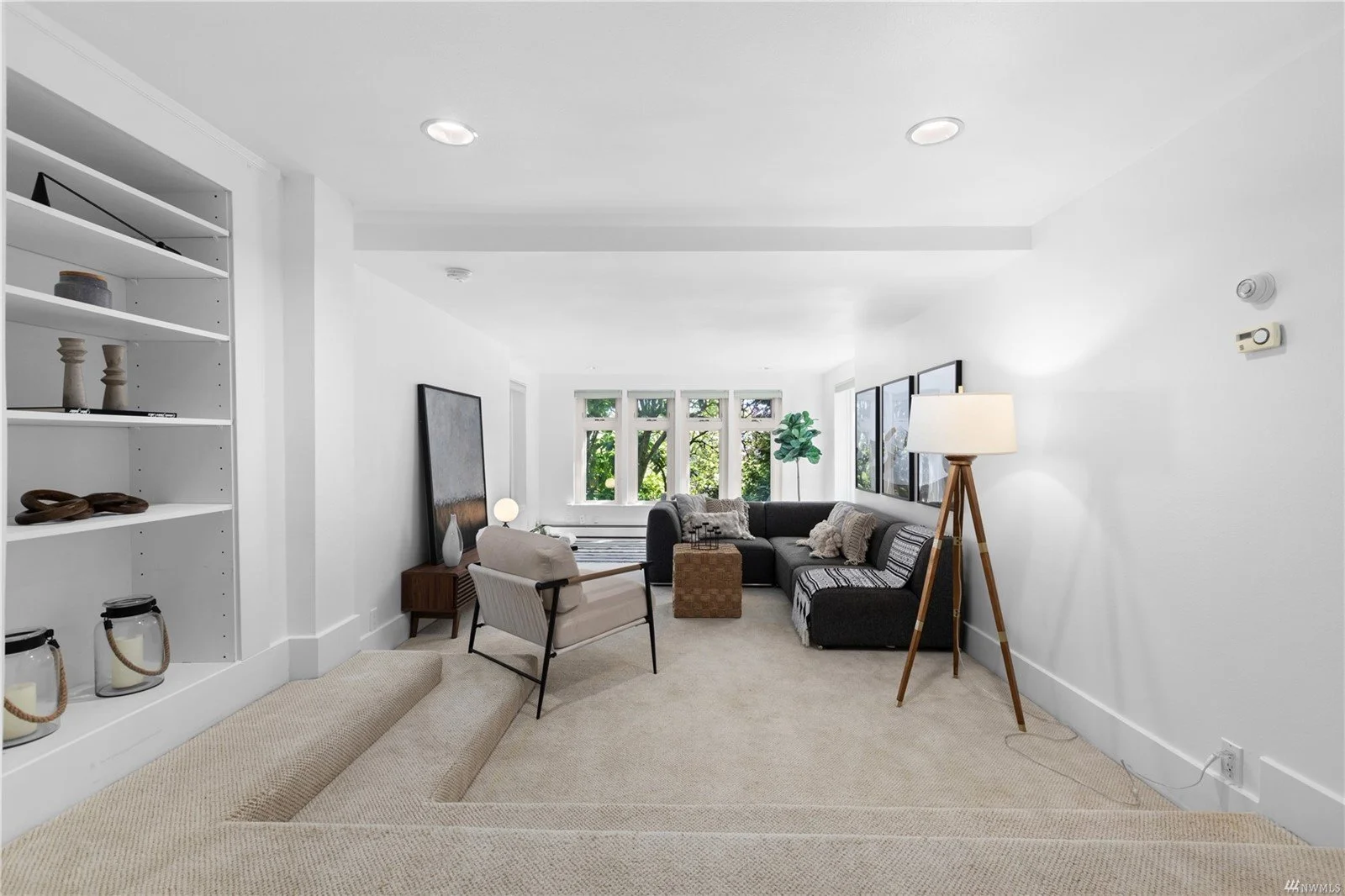 Bright living room with white walls, beige carpet, black sectional sofa, white armchair, standing lamp, framed artwork, and large window with trees outside.
