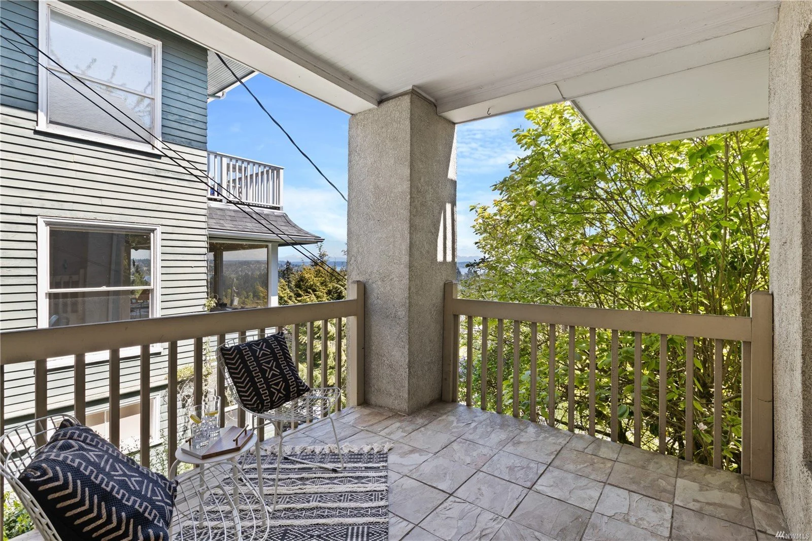 Balcony with tiled floor, two wire chairs with black and white cushions, small white side table with two glasses of water, and a colorful striped rug, overlooking a neighboring house with blue siding, a balcony, and a view of trees and a blue sky.