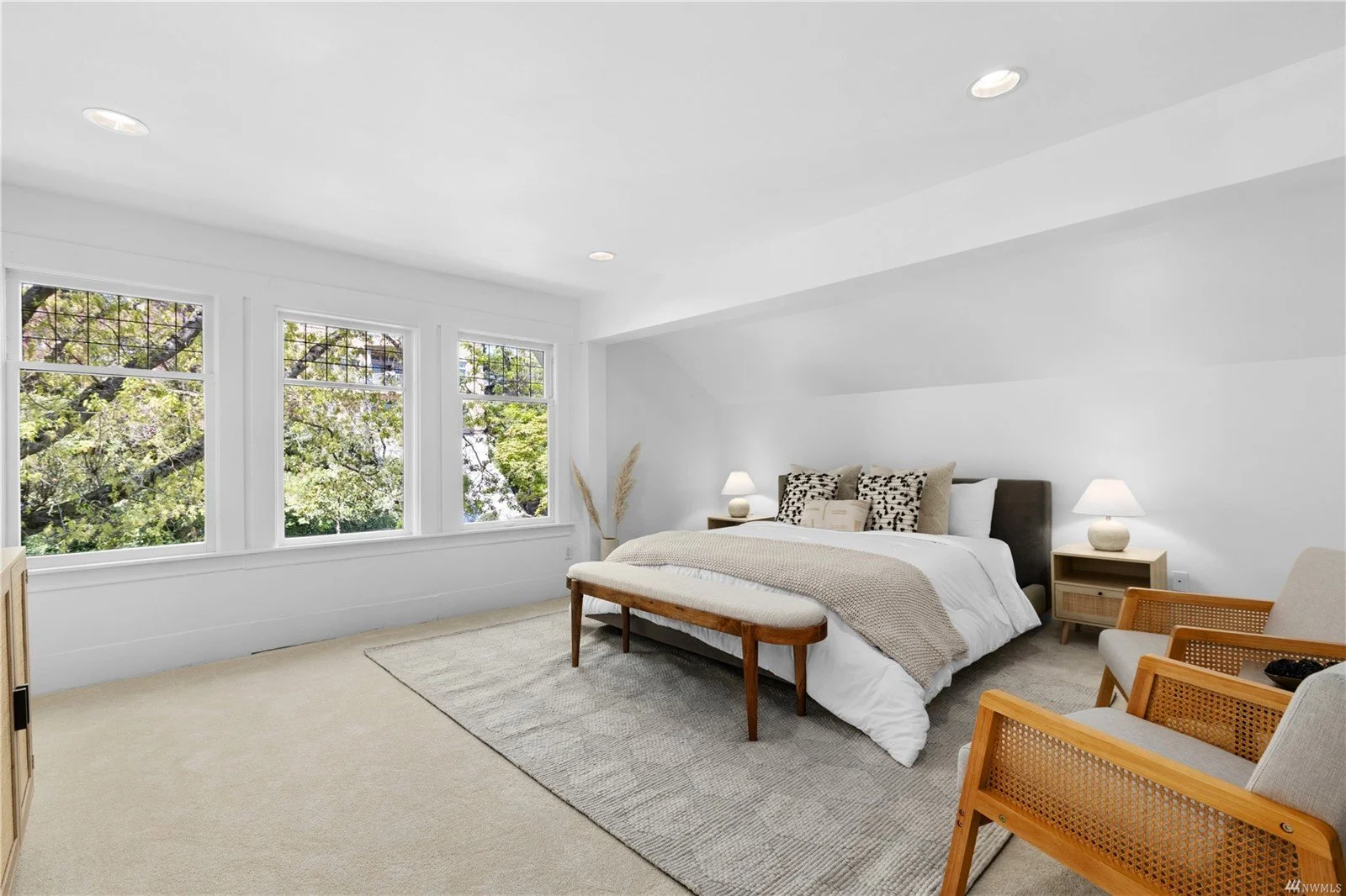 Bright bedroom with large windows, white walls, and a bed with beige and black pillows, a bench at the foot of the bed, two nightstands with lamps, and wooden chairs.