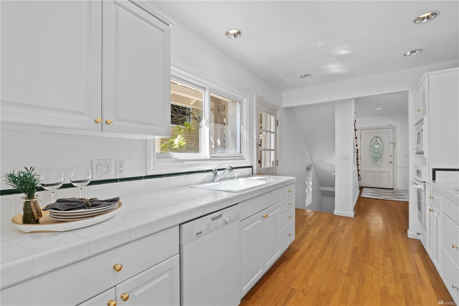 White kitchen with wooden floors, white cabinets with gold knobs, a white tile counter, and a sink beneath a window. Plants and glasses are on the counter, with a view of the entryway with a front door in the background.