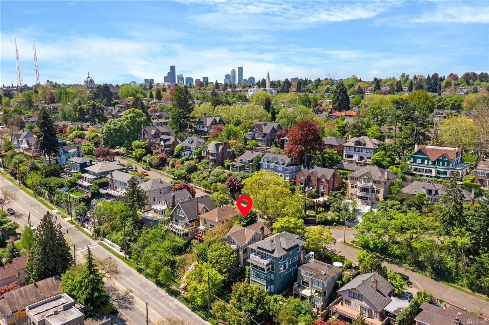 Aerial view of a residential neighborhood with numerous houses, trees, and greenery, with a city skyline in the background under a partly cloudy sky.