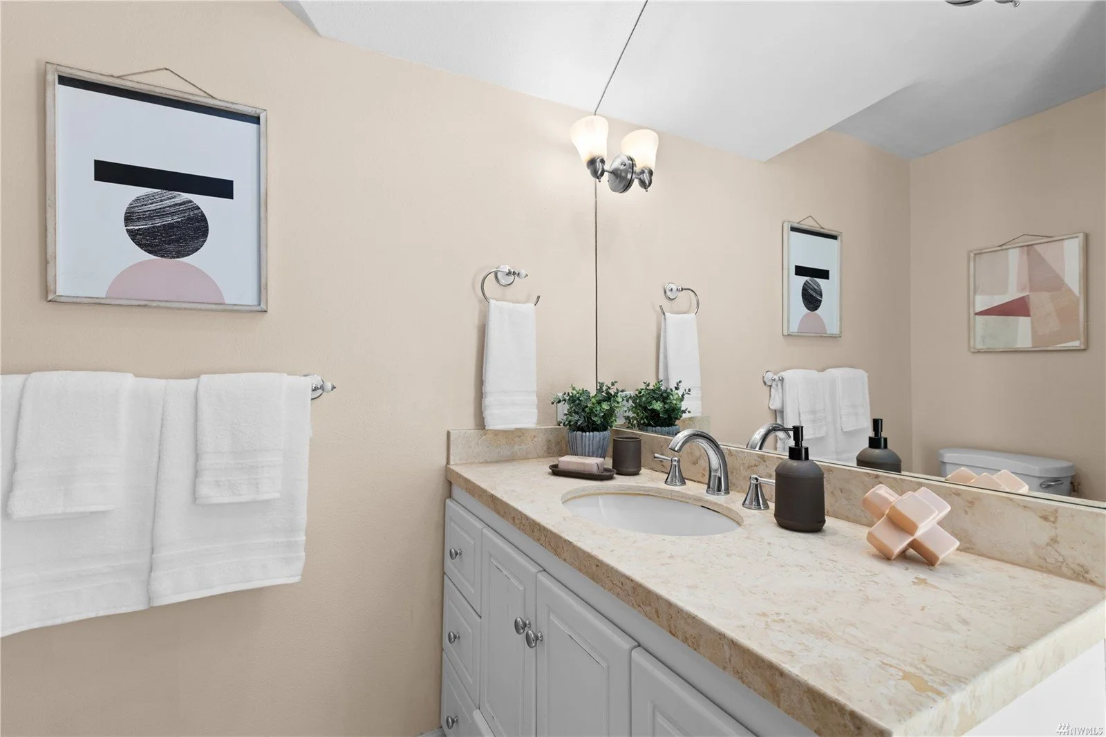 Bathroom with beige walls and white cabinetry. A large mirror above a light-colored marble countertop with a built-in sink, black soap dispensers, and small potted plants. Towel racks with white towels and framed abstract art on the walls. Ceiling li