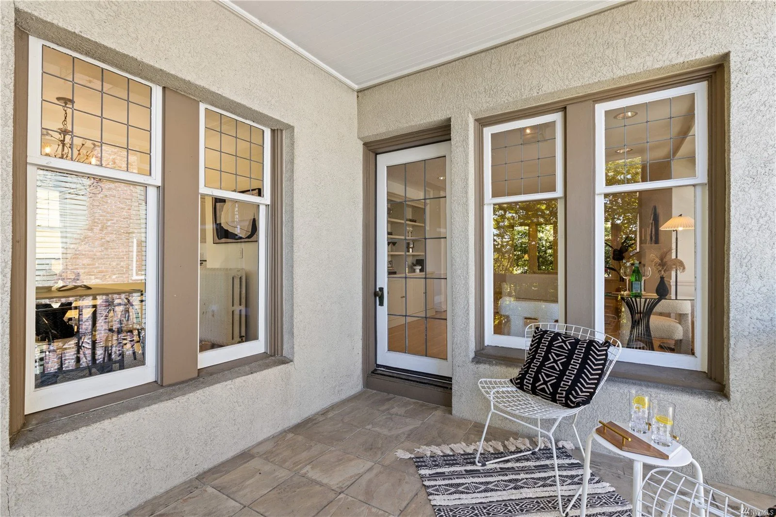 A small outdoor patio with beige stucco walls, large windows, a glass door, a white wire chair with a black and white patterned cushion, a small white side table with glasses of water and a tray, and a black metal table with a vase and plants inside 