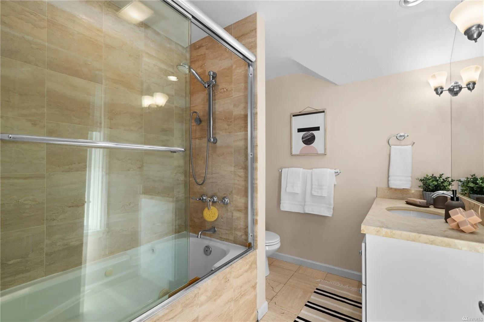 Bathroom with a glass-enclosed shower, a bathtub, a toilet, a beige countertop with sink, two potted plants, and artwork on the wall.