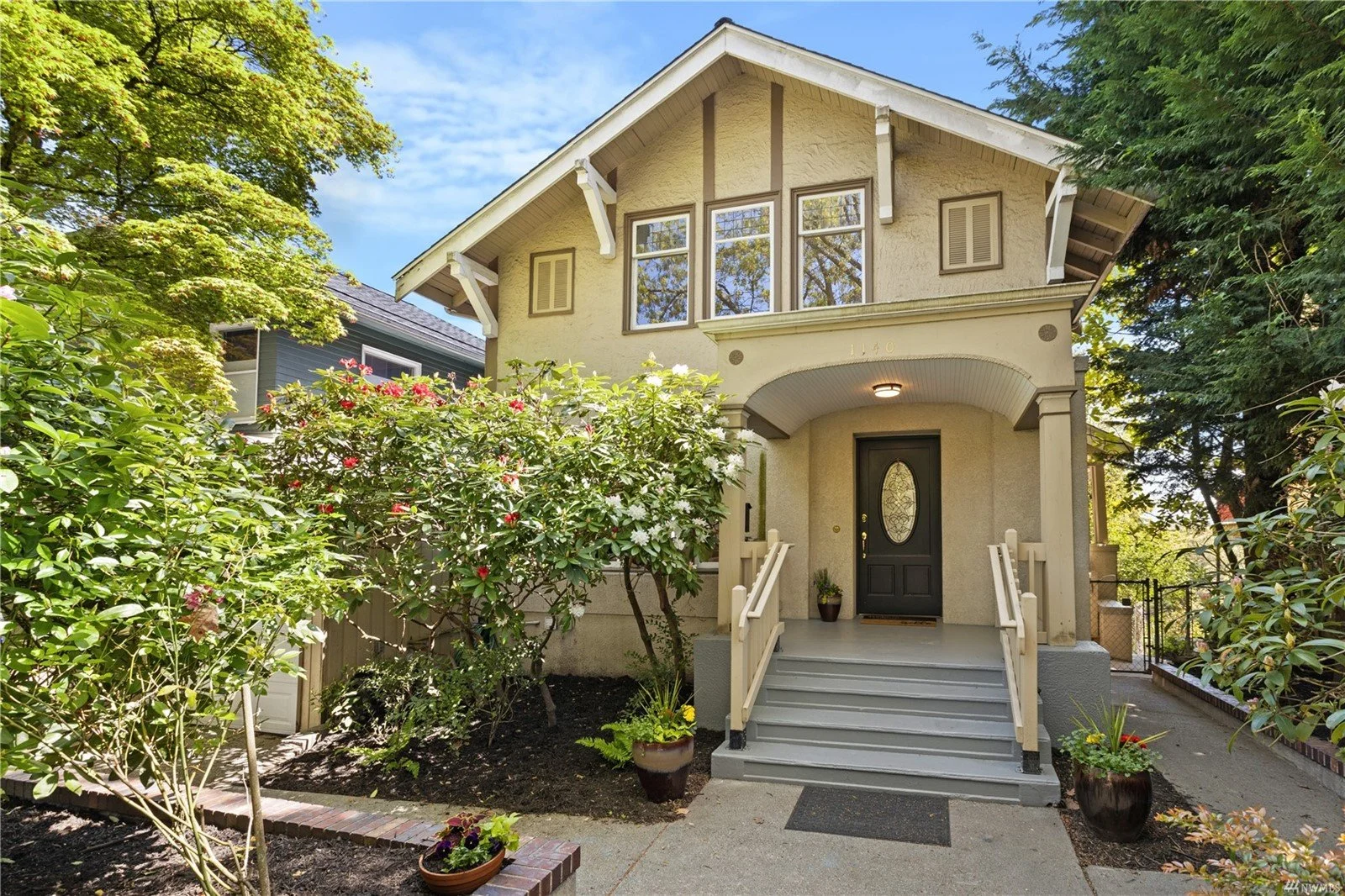 Front view of a two-story beige house with a dark door, surrounded by greenery and potted plants, with a small front porch and steps leading up to the entrance.