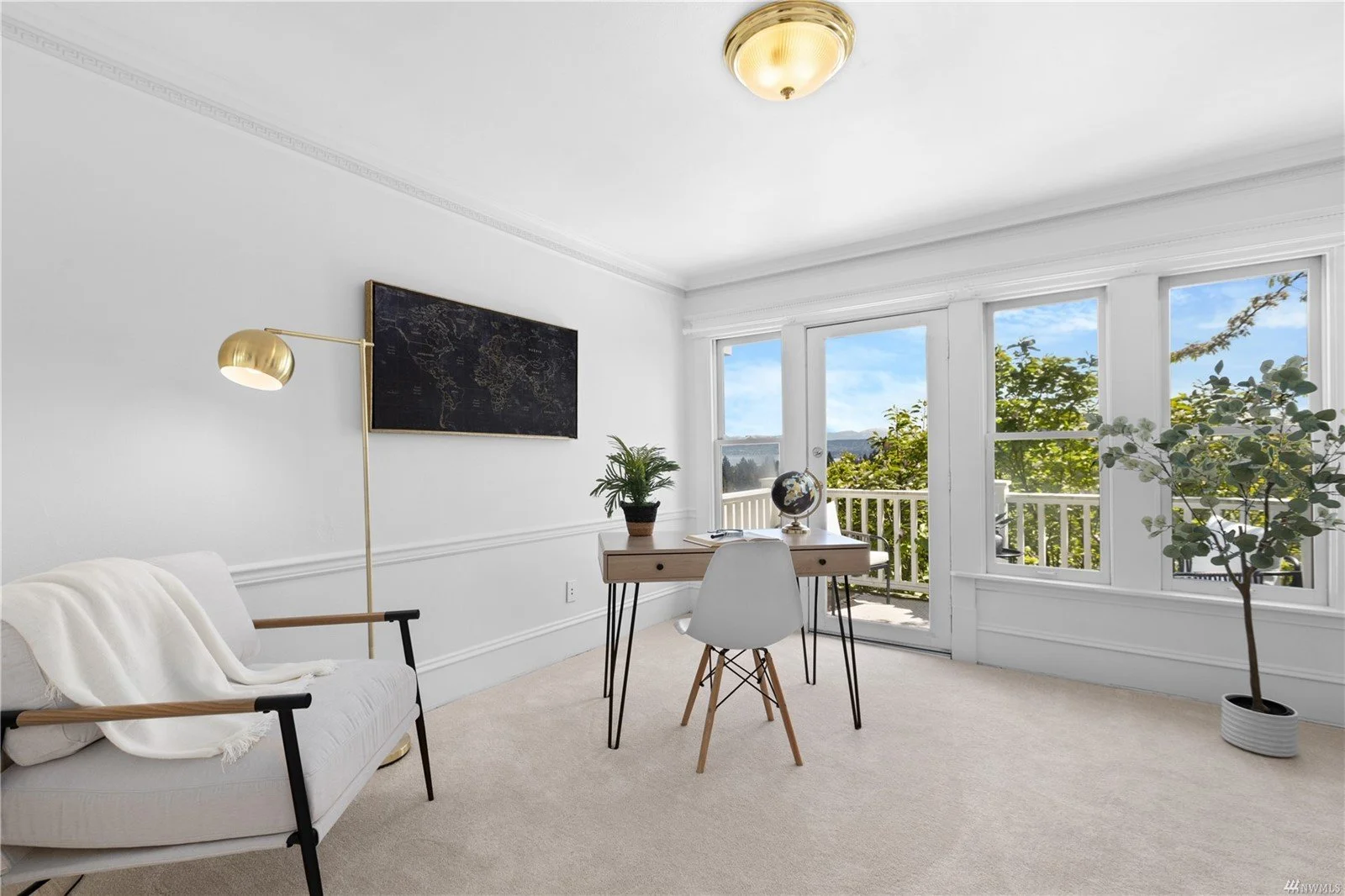 Bright living room with white walls, large windows with a view of trees, a small wooden desk with a globe and plants, a white chair, a beige sofa with a throw blanket, and a potted plant, illuminated by a ceiling light and a brass floor lamp.