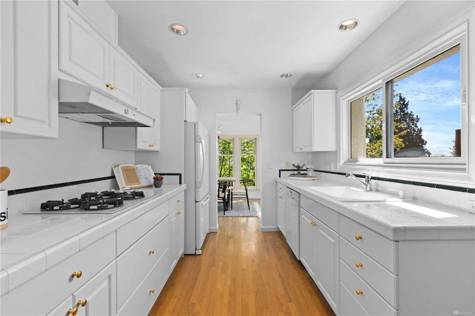 Bright kitchen with white cabinets, black countertops, a large window over the sink, and wood flooring, view into a dining area with a table and chairs, outside trees visible.