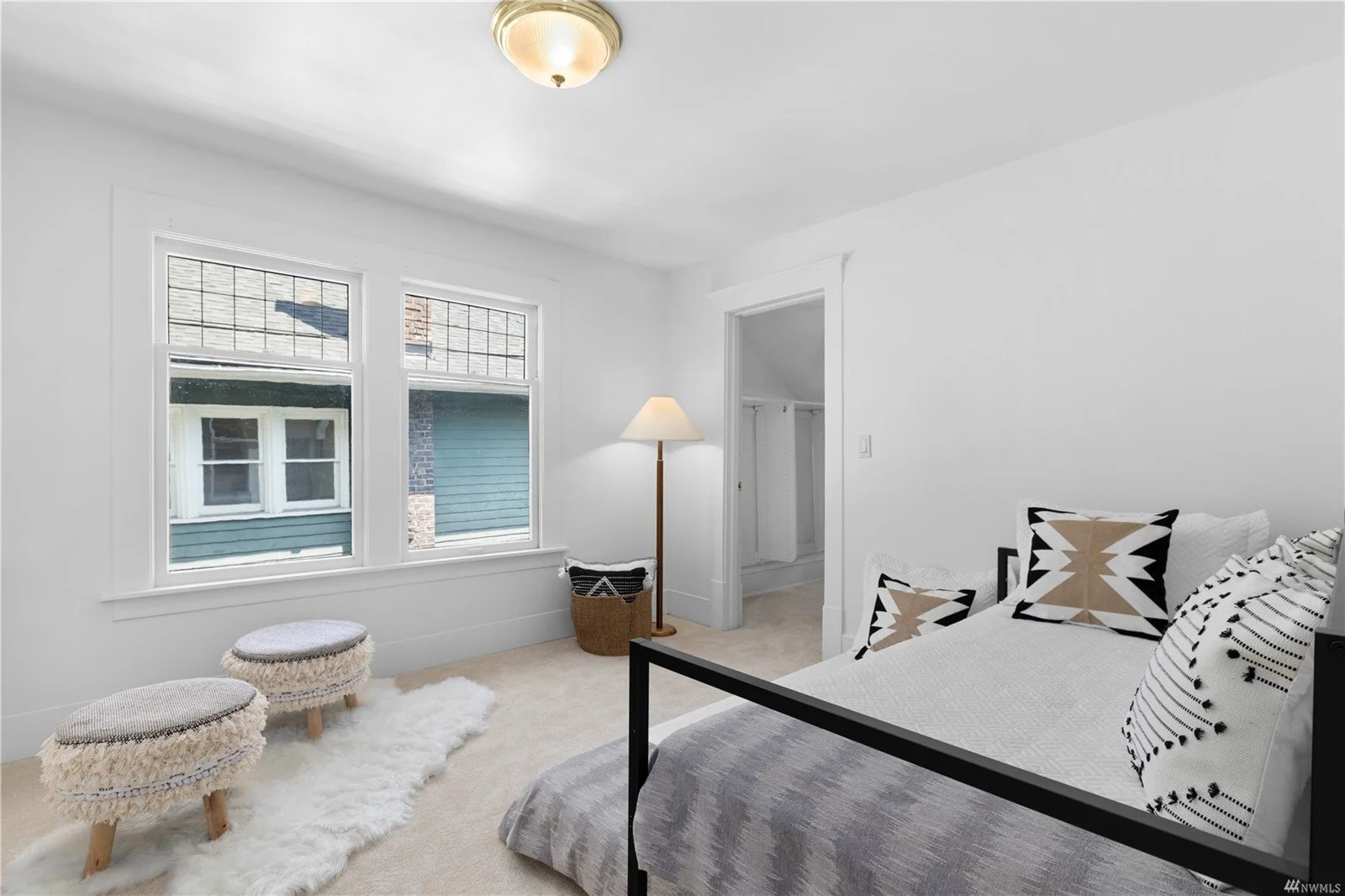 Simple bedroom with white walls, a bed with black frame, patterned pillows, a standing lamp, two small upholstered stools on a fluffy white rug, and two windows showing neighboring buildings.