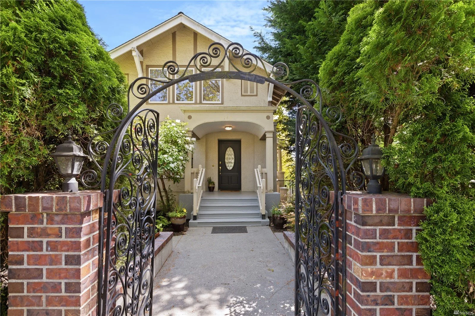 Front entrance of a two-story house with a black front door, steps, and a garden. The house is viewed through an ornate black wrought iron gate flanked by brick pillars with lanterns, surrounded by lush green bushes and trees.