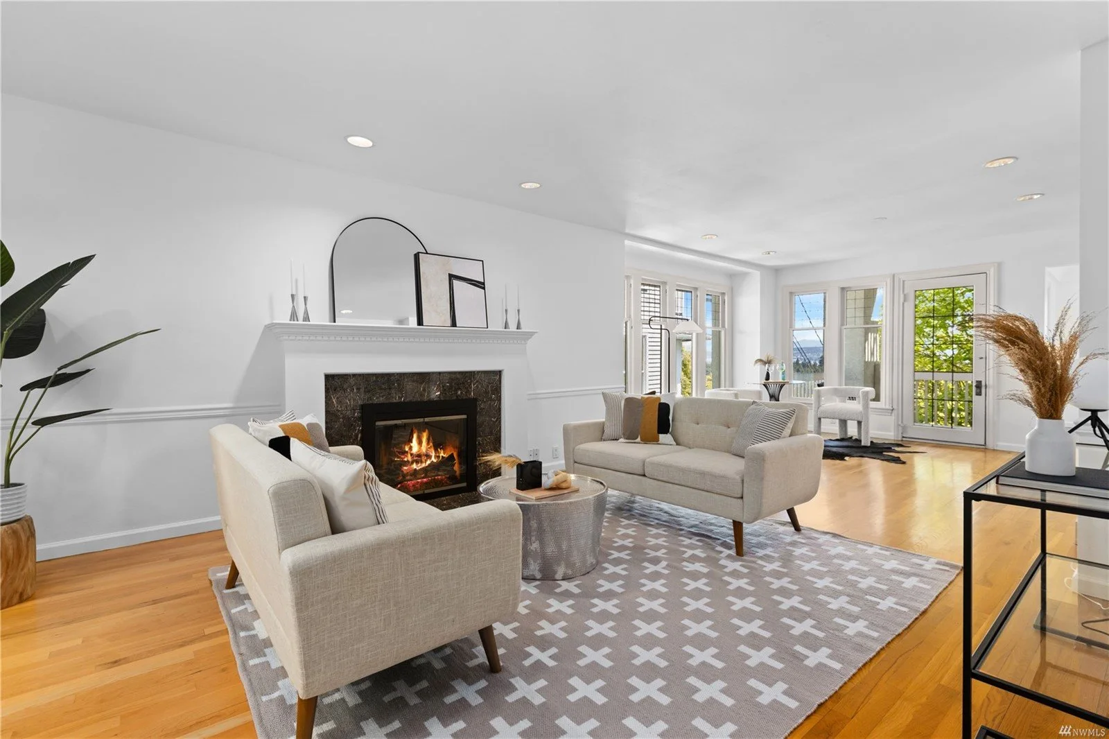 Bright living room with white walls, hardwood floors, a fireplace, and large windows with views of greenery outside. Contains beige sofas, a patterned rug, and decorative items.