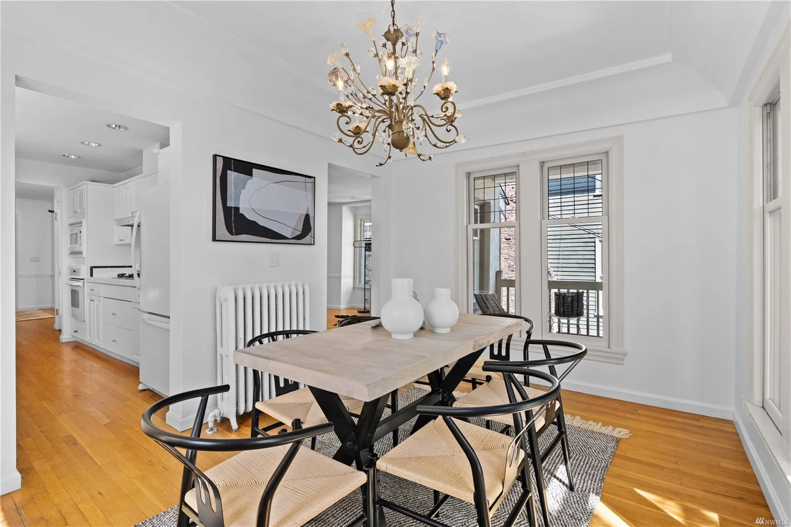 Dining room with a light wood table, black chairs, white vases, and an ornate chandelier. The room has white walls, large windows, and hardwood floors.