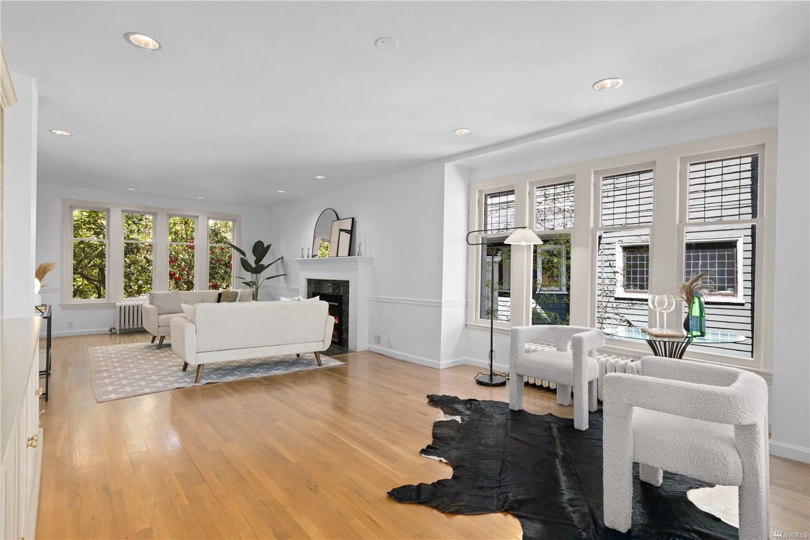 Bright living room with white walls, hardwood floors, multiple windows, and modern furniture including white sofas, a fireplace, and decorative plants.