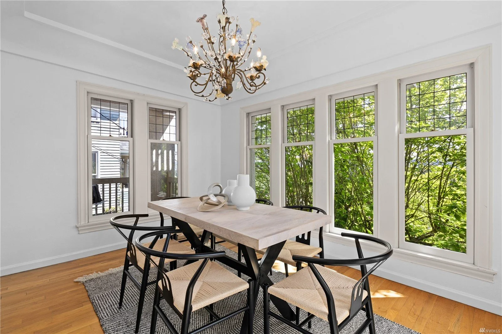 Bright dining room with white walls, large windows showing green foliage, wooden floor, a light-colored rectangular table with black chairs, white vases on the table, and a decorative chandelier hanging from the ceiling.