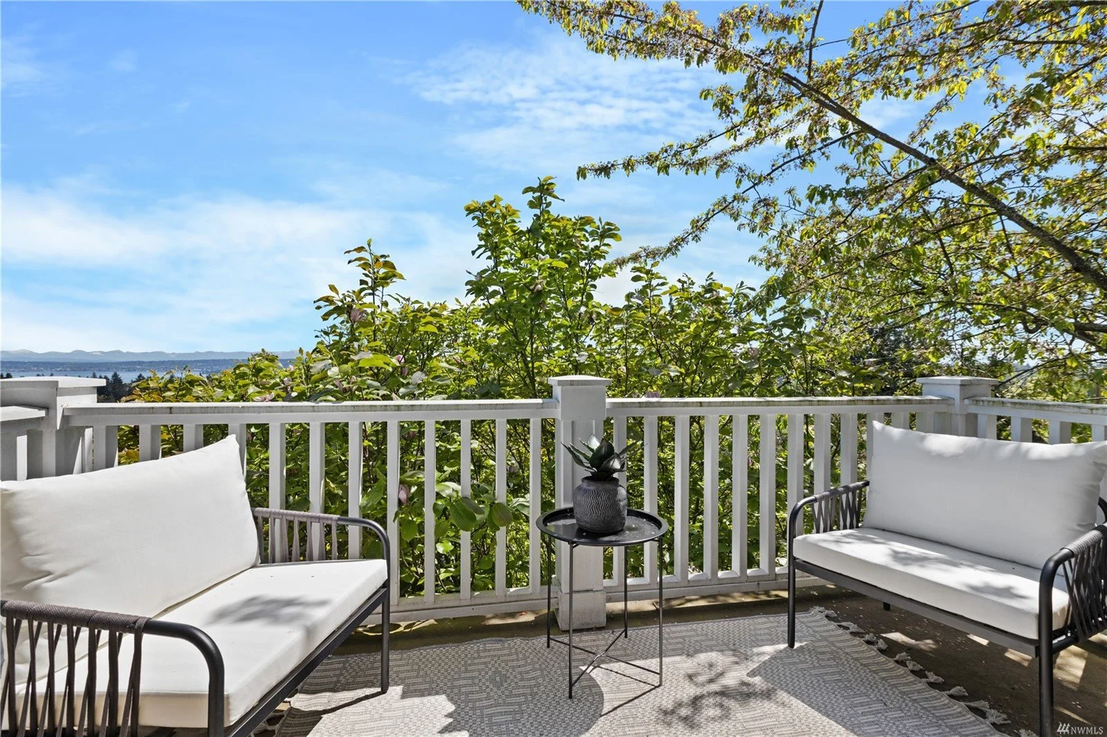 Outdoor balcony with white railing, two black metal chairs with white cushions, a small round black table with a dark plant in a vase, lush green trees, and a blue sky with some clouds in the background.