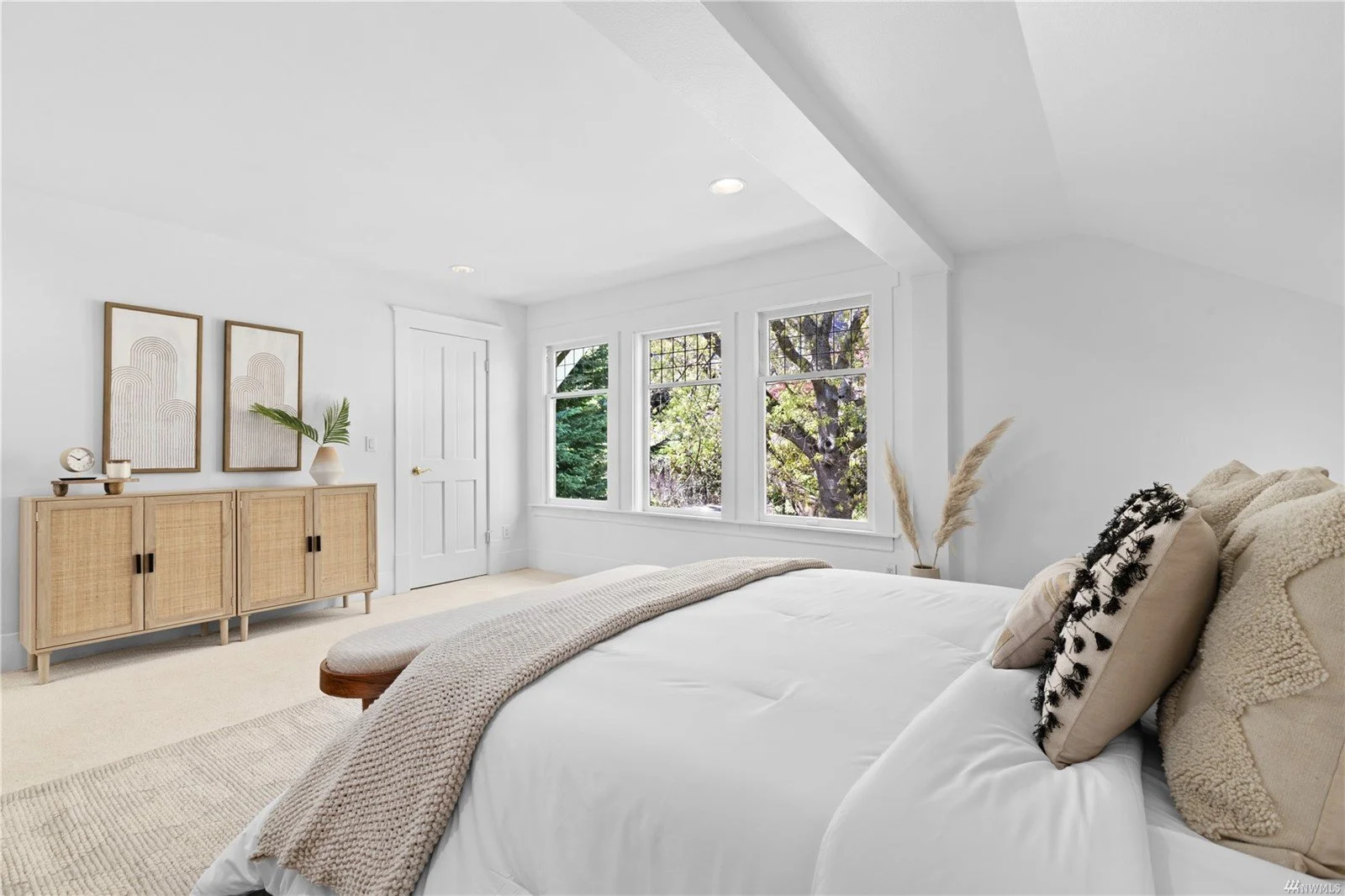 Bright, minimalist bedroom with a large bed, neutral bedding, four windows showing trees outside, a light wooden sideboard with decor, and neutral wall art.