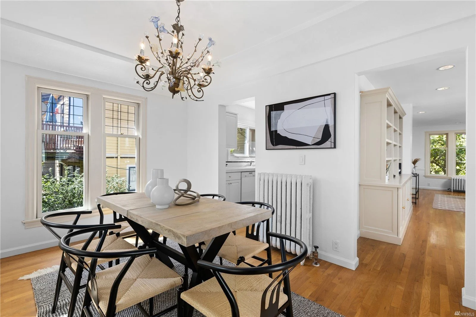 Dining room with a wooden table surrounded by black chairs with woven seats, white vases on the table, a chandelier hanging from the ceiling, large windows, and a living area with walnut flooring and second windows visible through an open doorway.